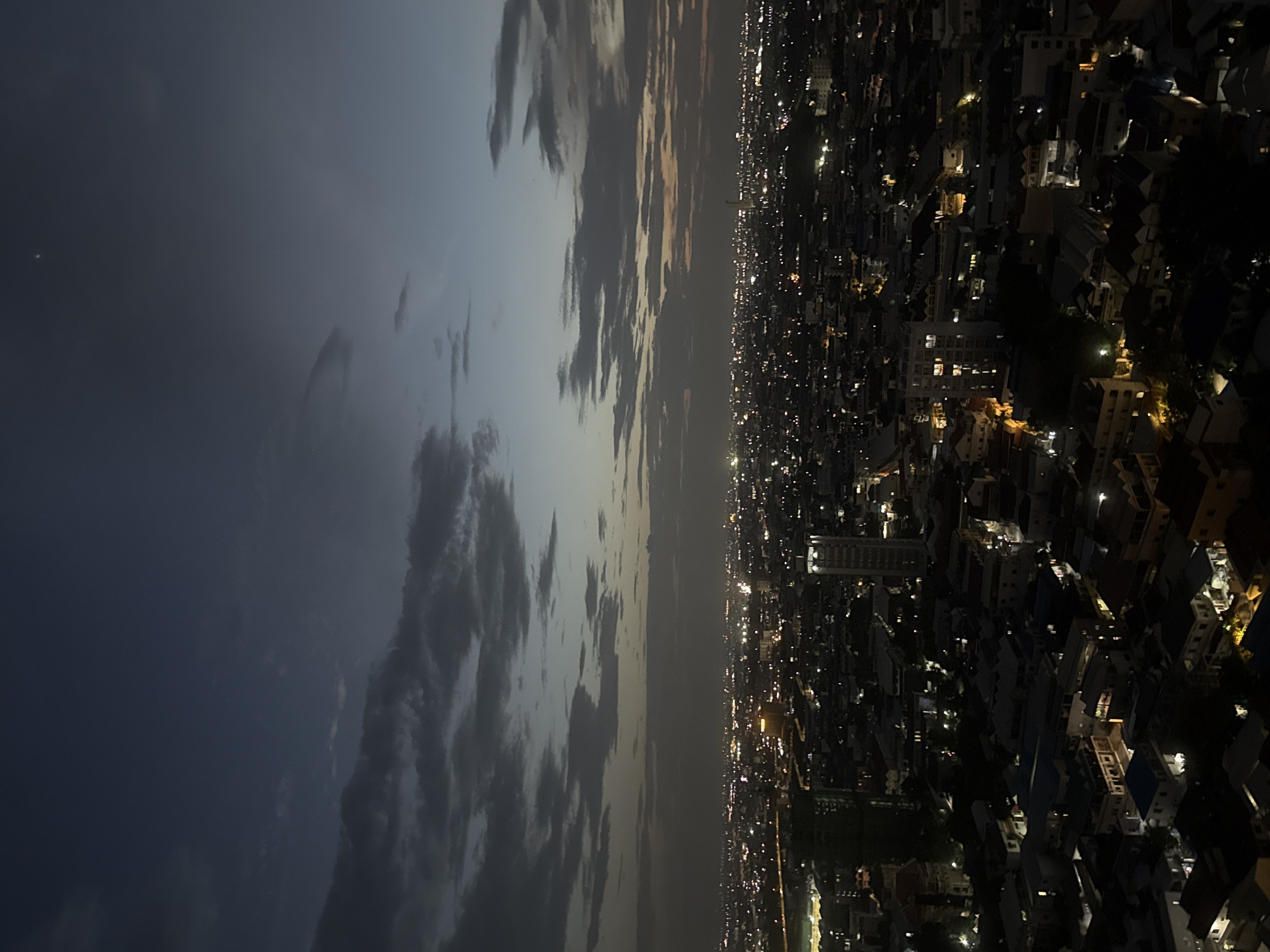 This is a high-angle, long-shot, dusk photograph of a sprawling city illuminated by artificial lights. The sky is a deep, dark blue, transitioning to a lighter grey near the horizon, with scattered, wispy clouds. A single bright point, possibly a planet or star, is visible in the upper right portion of the sky.
The city below is a dense mosaic of buildings of varying heights, with countless lights glinting from windows and streets. In the foreground, darker, lower-rise structures with visible rooftops dominate the scene. As the view recedes into the distance, taller buildings, including distinct skyscrapers and a building with a prominent, square-shaped top, become more apparent. The overall impression is one of urban density and activity.
While the image captures a broad view of the city, specific details about people, actions, or interactions are not discernible due to the distance and low light conditions. The photograph is a forensic depiction of the urban landscape at twilight. Based on the provided context, the scene is located in Khan Mean Chey, Cambodia.
