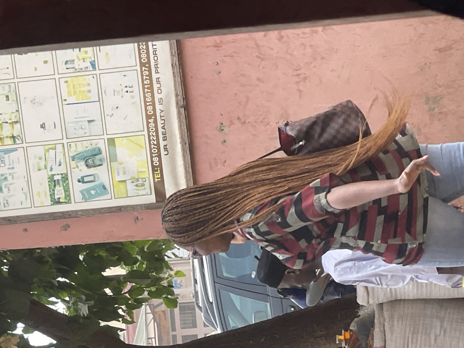 The image captures an outdoor street scene in Samaru, Nigeria, during daylight. A woman with dark skin is prominently featured in the mid-ground, walking from right to left. She wears eyeglasses, a patterned short-sleeved top with red, black, and greenish-gray blocks, and light blue jeans. Her long, light brown braided hair extends below her waist, appearing to be in motion. She carries a brown checkered shoulder bag with a red interior lining on her left shoulder. Her head is turned to the left, revealing a smiling or laughing expression. Behind her and to the left, partially obscured by a tree trunk, stands a man with dark skin wearing a black skullcap and a white shirt. To the right, a faded pink wall displays a white rectangular advertisement sign for beauty products, featuring images of items like CeraVe, Nigerian phone numbers, and the slogan "UR BEAUTY IS OUR PRIORITY." In the left background, a dark-colored SUV is visible, and below the tree, white sacks and small containers with amber liquid indicate a street vendor's display.