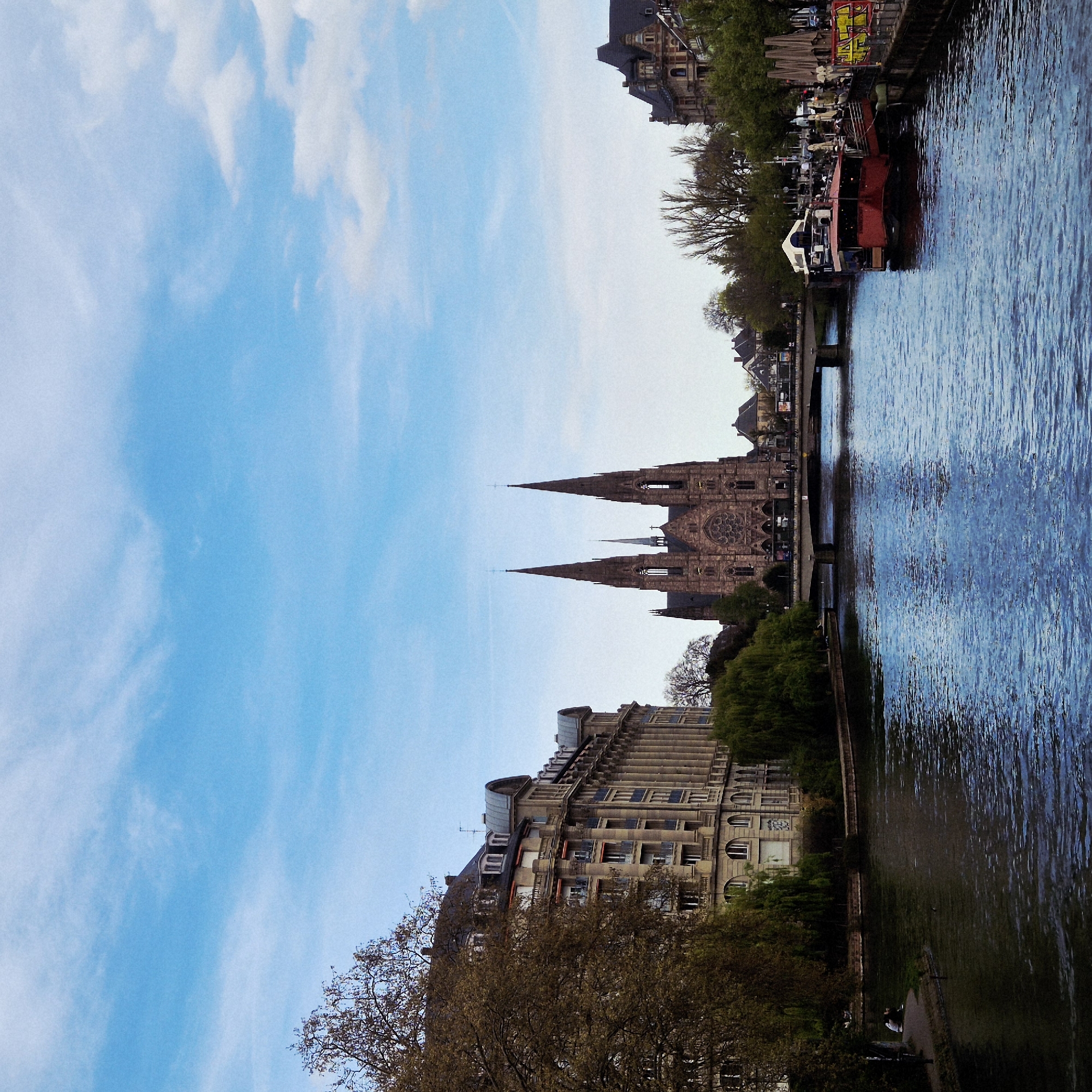 The photograph shows the Strasbourg Cathedral in France, seen from across the Ill River. The cathedral, a large Gothic structure, dominates the center of the frame with its distinctive spires. The Ill River is on the right side of the image, with reflections of the surrounding buildings and sky. On the riverbank, there are buildings and trees, as well as a tour boat. The sky is visible on the left, with scattered clouds.
