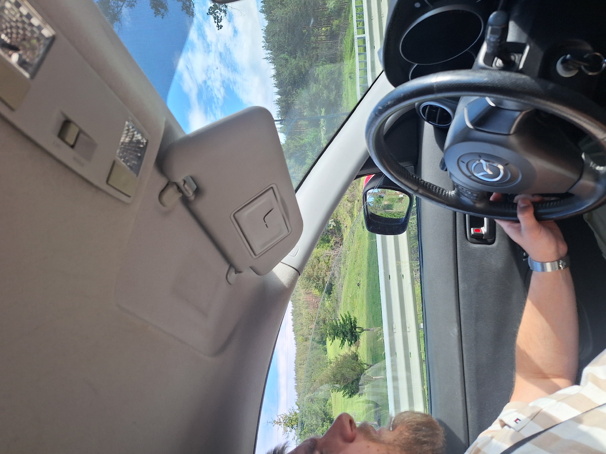 The image shows a driver seated in a Mazda car. The driver's hands are on the steering wheel and their forearm is visible, adorned with a wristwatch. The car's interior features a sun visor, dome lights, and a rearview mirror. The driver appears to be on a road near Belchin, Bulgaria, suggested by the landscape visible through the window. The environment outside the car includes a green, grassy terrain and forested areas under a sky with scattered clouds. The image is taken from a slightly skewed perspective, looking towards the front and side of the car's interior.