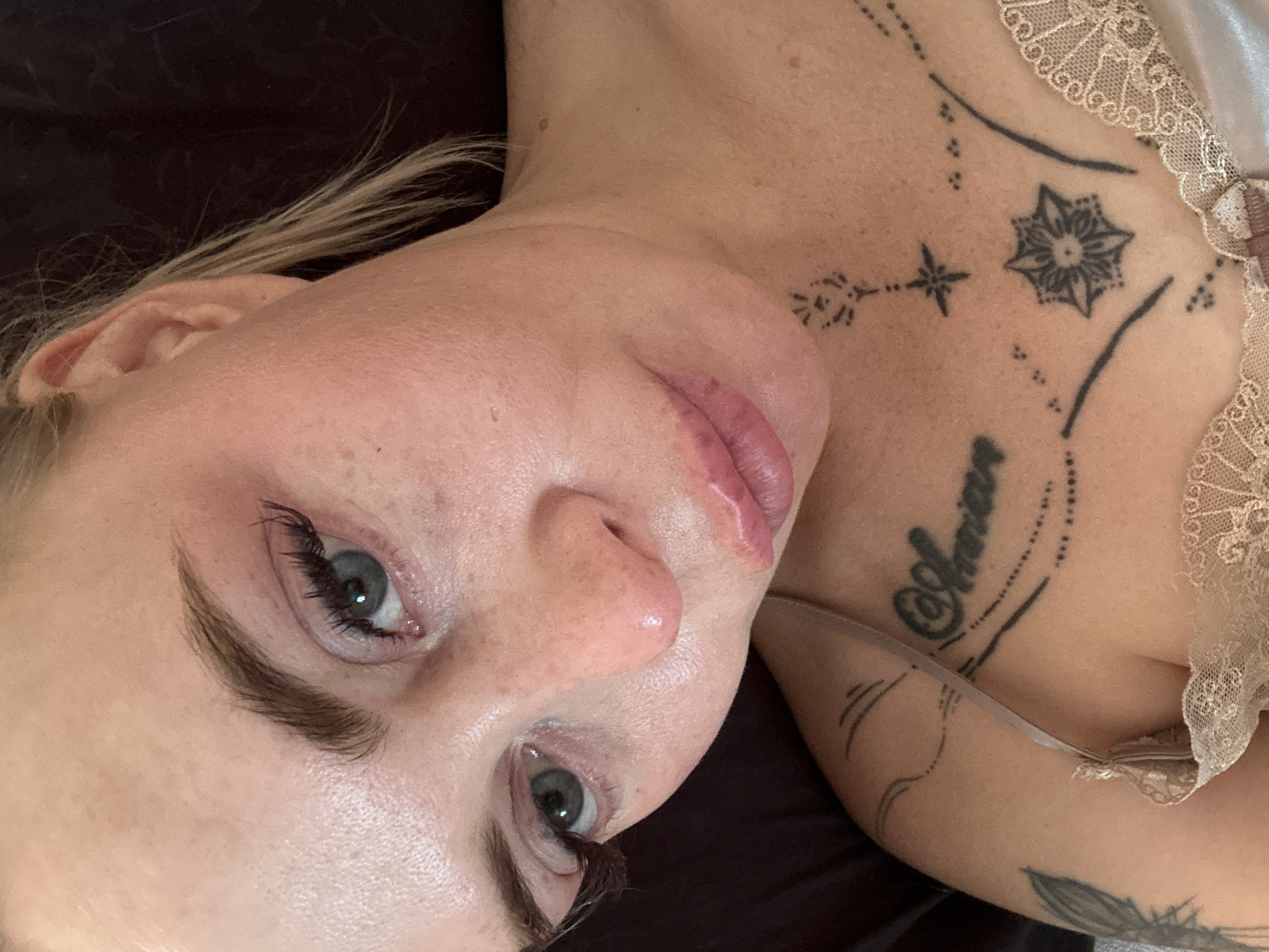 A close-up, overhead shot shows a woman with fair skin, blonde hair pulled back, and light blue eyes. She is wearing a beige lace bralette and has visible tattoos on her chest and shoulder. The tattoos include script, a star design, and floral patterns. The woman's makeup includes mascara and lip gloss. The background is dark and appears to be a pillow or blanket. The scene is indoors, with no discernible city or location information.