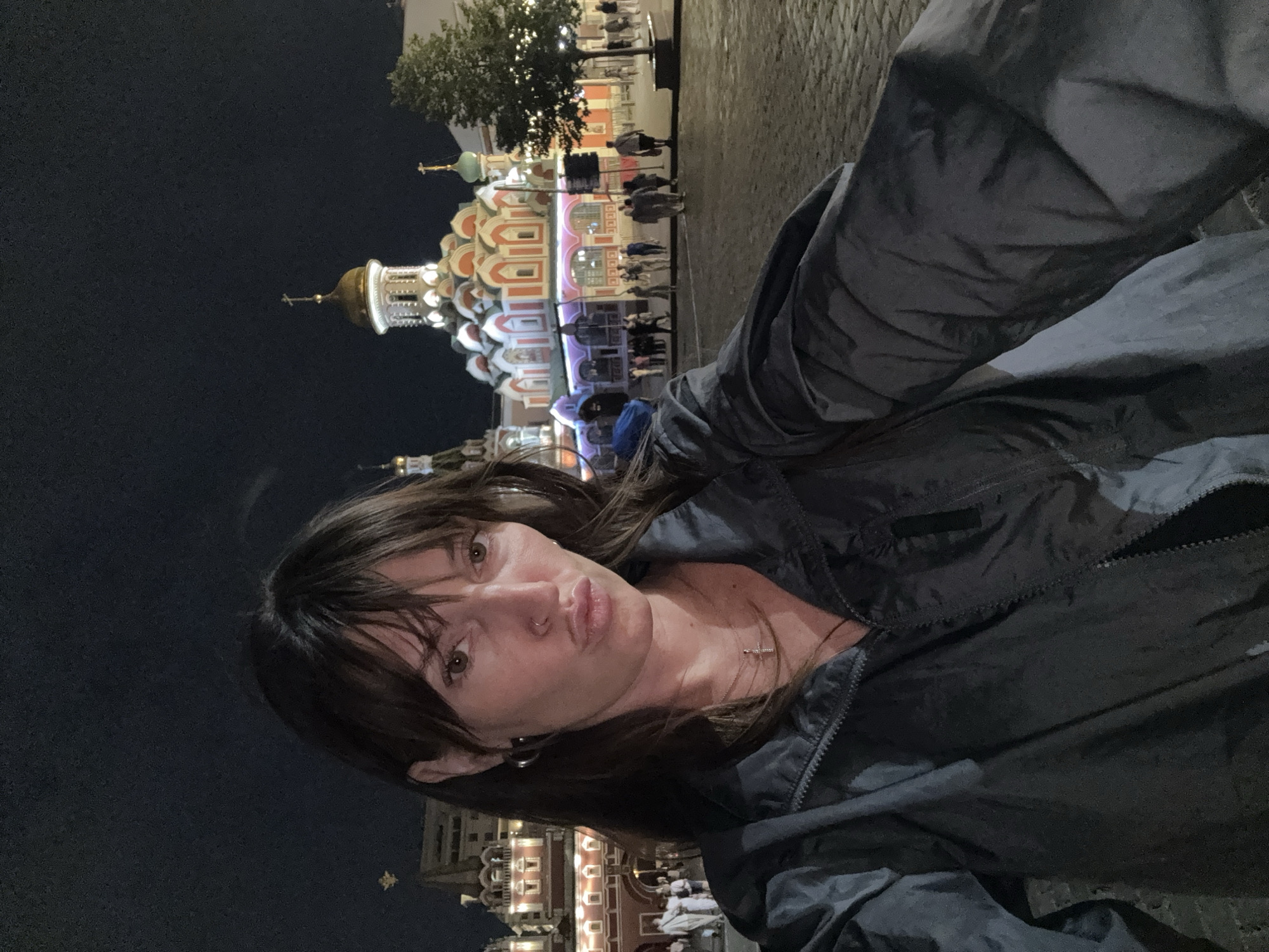 A medium shot selfie captures a woman with brown hair and bangs, wearing a dark jacket, a cross necklace, and hoop earrings. Her lips are pursed. In the background, a city square at night is illuminated by lights. A prominent building with a red facade and a white and gold domed structure with crosses are visible. People are scattered throughout the square, and a tree stands to the right. The ground is paved with cobblestones.