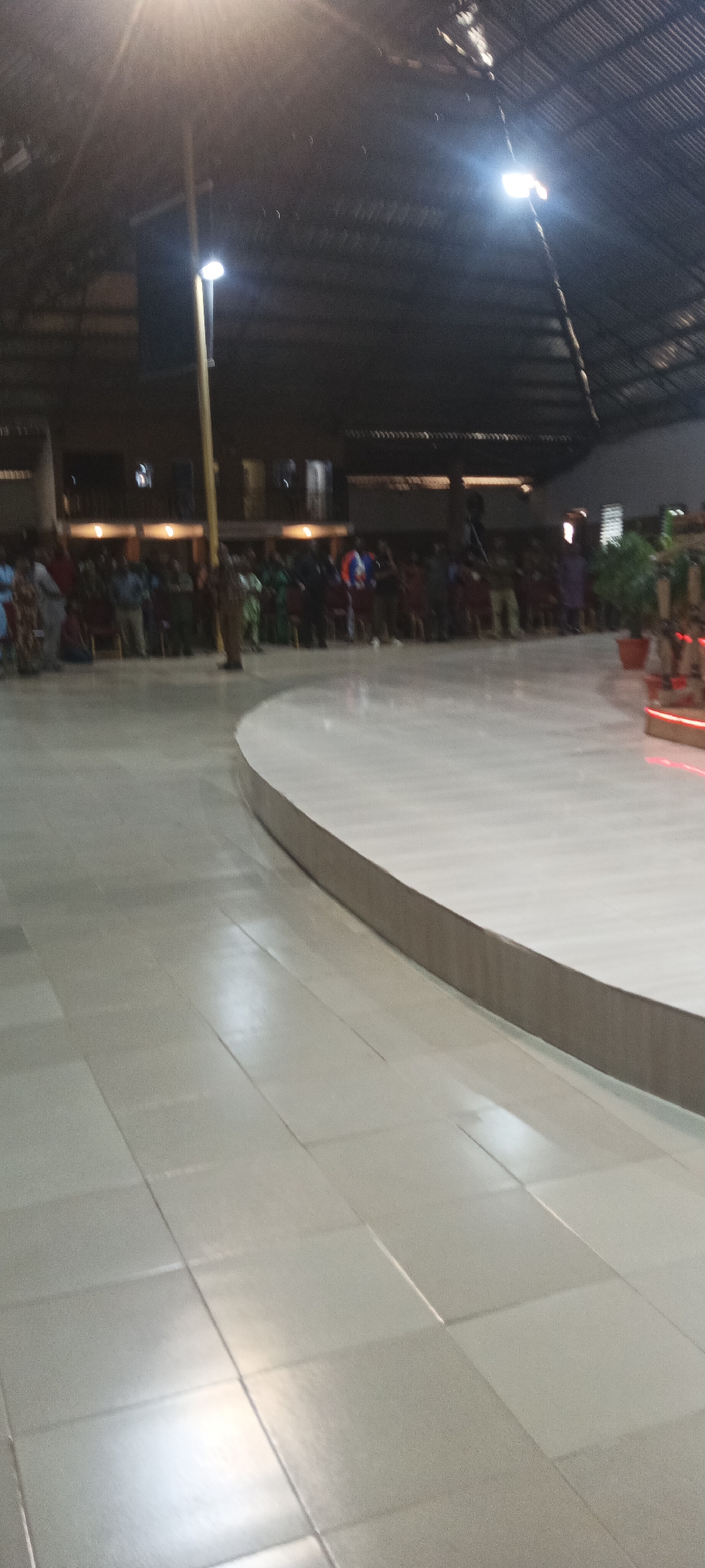 The image captures a wide-angle view of an indoor gathering, possibly at a church or community hall, situated on Rukuba Road, Nigeria. The scene is dimly lit, with overhead lights casting a warm glow on the corrugated metal roof and a long, central yellow pole. In the background, a crowd of people stands in rows, facing what appears to be a stage or focal point, though the details are blurred. Some individuals are standing, while others are seated on chairs. The foreground shows a polished, light-colored tiled floor that curves to the right, forming a raised platform. The overall impression is one of a communal event taking place in a large, enclosed space.