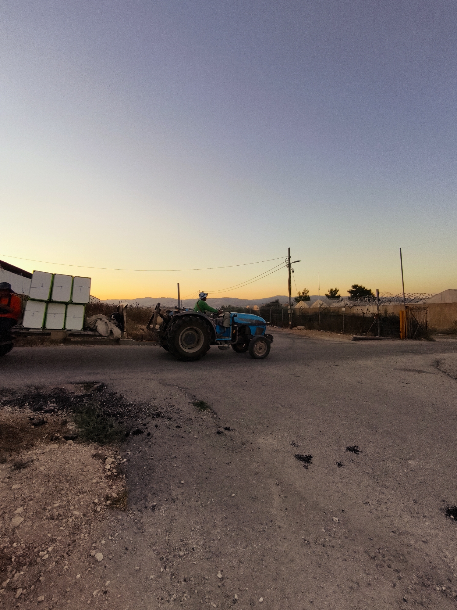 The image shows a blue tractor pulling a trailer loaded with white, square-shaped objects, possibly produce packaging, on a paved road. A person wearing a green shirt and a light-colored helmet is operating the tractor. The trailer is attached to a dark-colored vehicle on the left. In the background, there are buildings, fences, and vegetation under a clear sky with the colors of sunset. The scene appears to be in an agricultural or industrial area, possibly in the city of Lachish Regional Council, Israel, given the presence of greenhouses in the background. The ground is a mix of asphalt and dirt. The overall lighting suggests it is either sunrise or sunset.