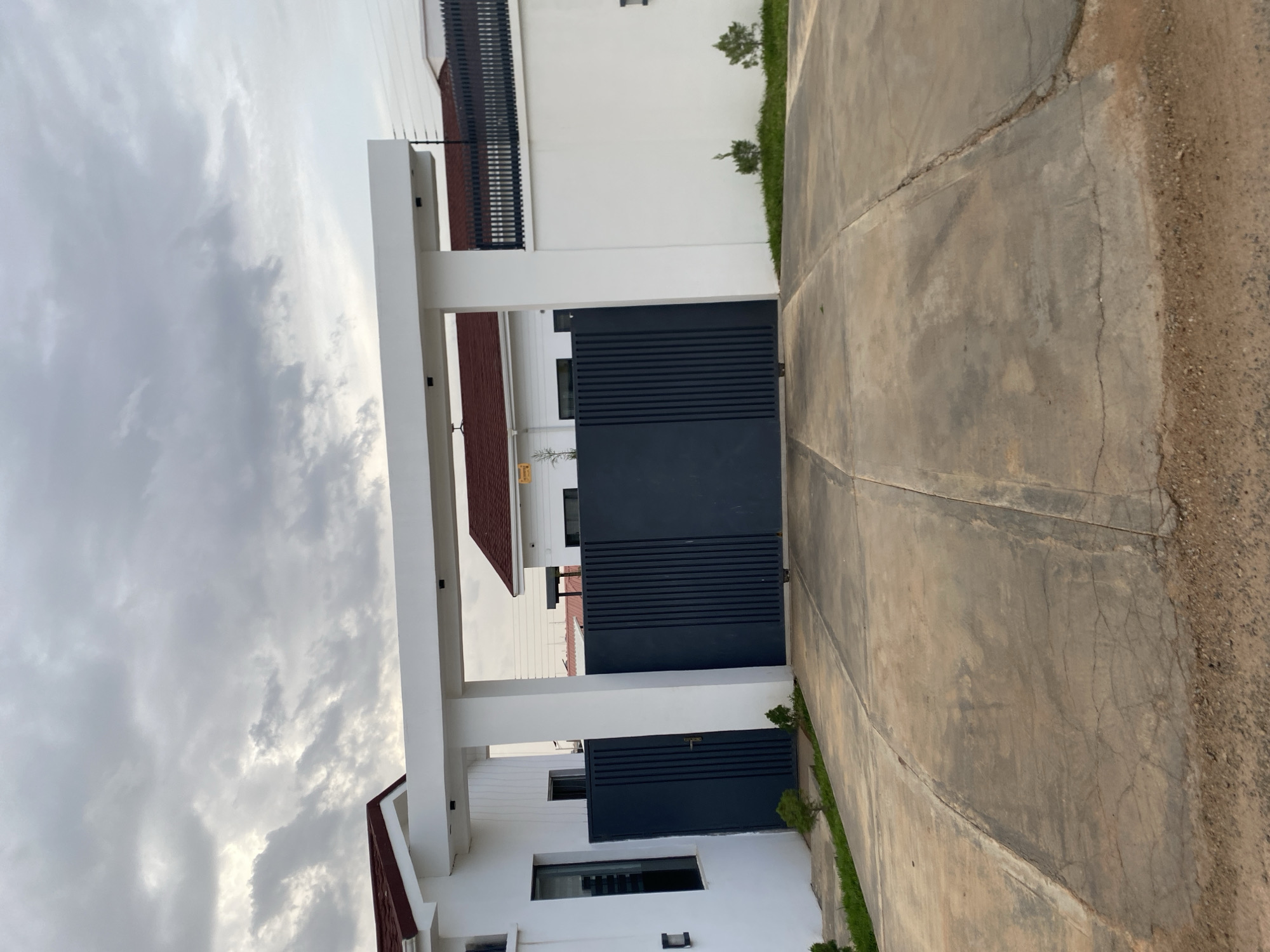 This is a frontal, eye-level photograph of a modern house entrance in Barikin Jirga Summa, Nigeria. The scene is dominated by a large, dark grey metal gate, which is closed. The gate is flanked by white pillars and a white horizontal beam, forming an entryway. To the left of the gate is a section of a white house with a dark-framed window. To the right, a white wall topped with a black security fence extends into the background. The roof of the house is visible above the entryway and along the white wall, featuring a reddish-brown tile. The ground in front of the gate is a concrete driveway, showing signs of wear and cracking, with sparse green grass and small plants growing along the edges. The sky is overcast with heavy, grey clouds.