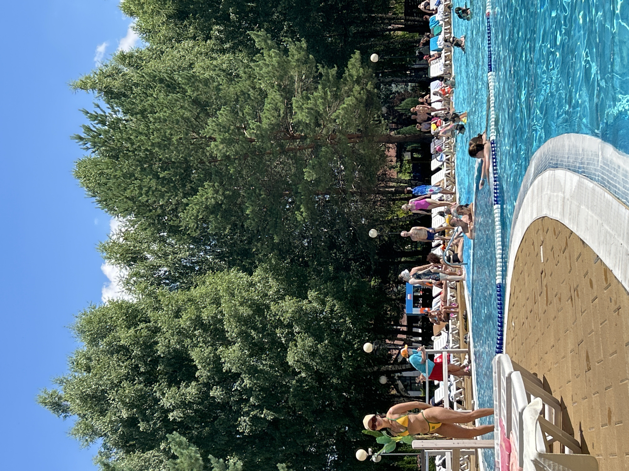 This image depicts a crowded outdoor swimming pool on a sunny day. The pool is surrounded by lounge chairs and trees. People are seen swimming, sunbathing, and socializing.
Without specific visual cues or landmarks, it is impossible to definitively identify the location as Togliatti, Russia. The scene is typical of many outdoor swimming pool facilities in various locations.