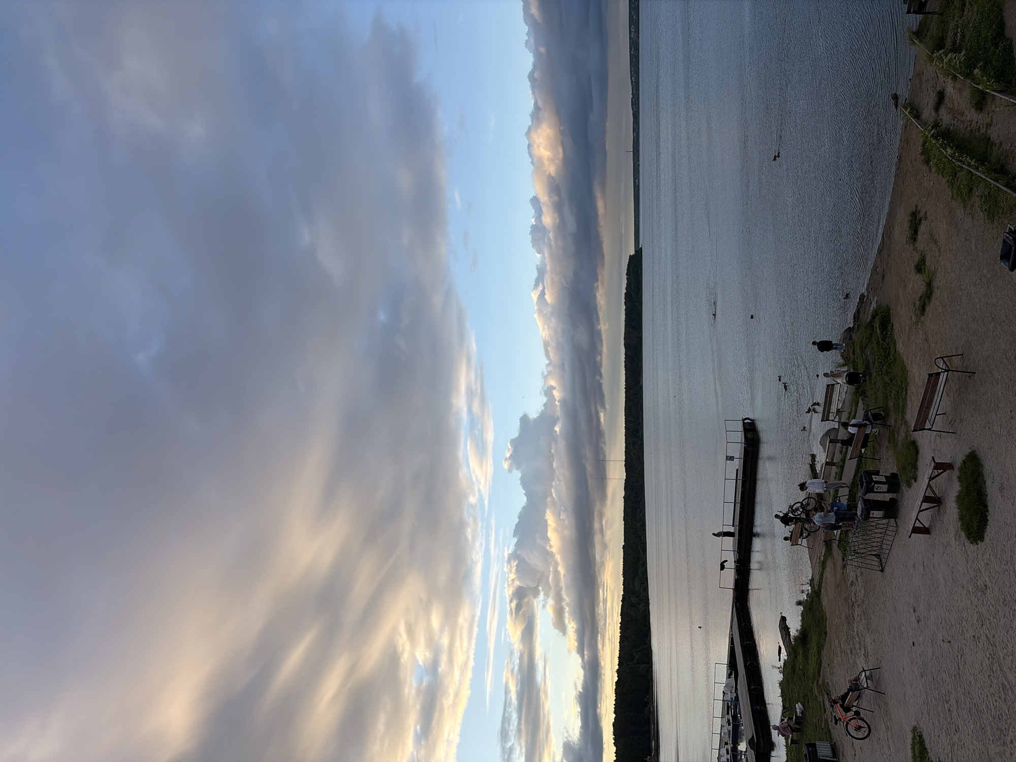 The image depicts a waterfront scene with a wide body of water, likely a lake or river, stretching across the frame. The sky is filled with dramatic clouds, tinged with warm light from what appears to be a sunset or sunrise. A dense line of trees borders the far bank of the water. In the foreground, a sandy and grassy area features a wooden pier with several people standing on it. Further inland, there are benches, bicycles, and more individuals engaged in various activities. The overall atmosphere suggests a public recreational area. This scene is located at the Shartash Stone Tents Nature Reserve in Yekaterinburg, Russia, a popular spot for outdoor activities and enjoying the natural beauty of the region.