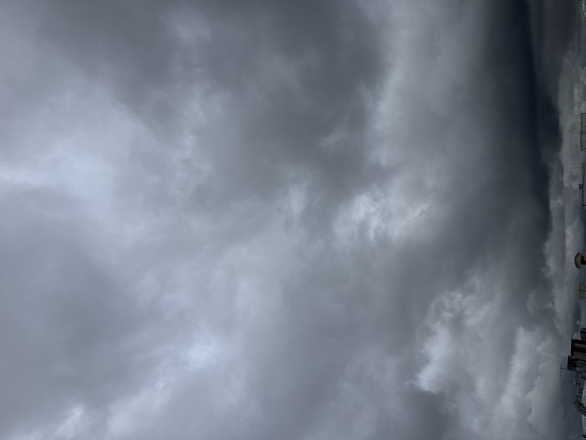 The image depicts a sky filled with heavy, dark storm clouds over a cityscape. The clouds are a uniform, ominous grey, suggesting impending rain or a thunderstorm. In the lower portion of the image, the tops of buildings are visible, indicating an urban environment. These buildings are mostly dark and unadorned, contributing to the somber mood of the scene. The overall impression is one of a brooding, overcast day in a city. No people are visible in the image, and no specific actions or interactions are captured. The location is identified as sovkhoza imeni Lenina, Russia, placing this stormy sky within that geographical context.