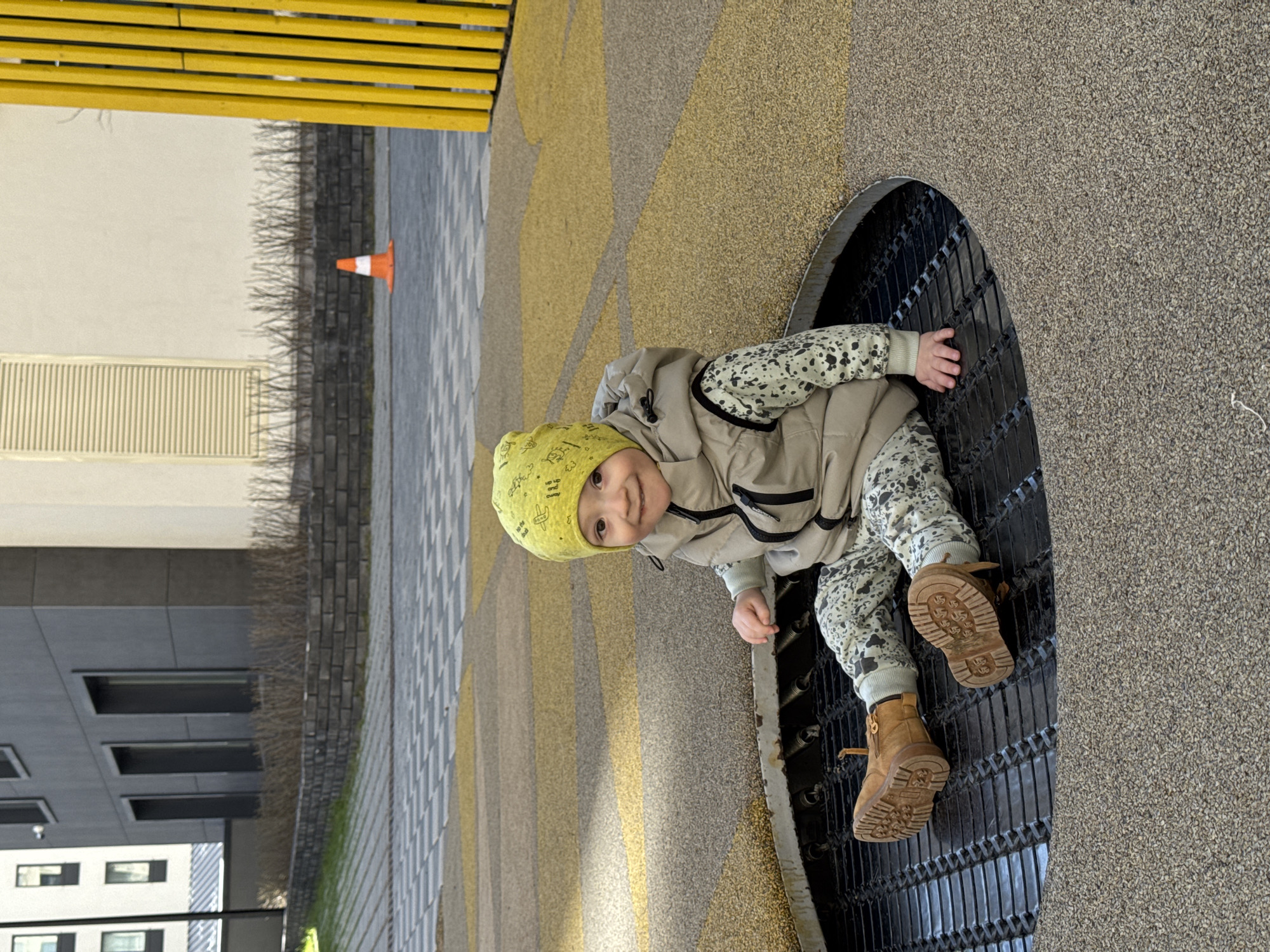 A child wearing a yellow hat, a vest, patterned clothing, and brown boots sits within a circular metal grate on a textured, geometrically patterned ground in what appears to be a modern urban setting. An orange cone is visible in the background, near a building with rectangular windows. The location is Izhevsk, Russia.
