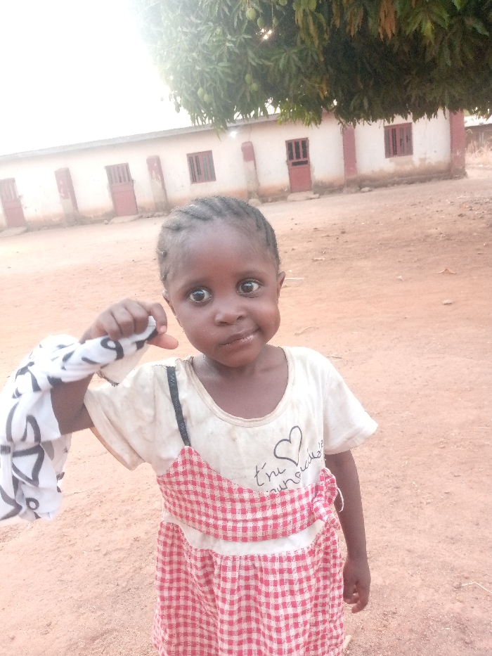 An outdoor scene in Agban, Nigeria, depicts a young child standing on reddish-brown, dusty earth, looking directly at the camera. The child has dark skin and short, dark braided hair. They wear a soiled, light-colored short-sleeved t-shirt with a black heart graphic and partially visible French text ("t'es m'a merveille") over a red and white checkered dress or pinafore, one black strap visible on the left shoulder. The child's left hand is raised, clutching a crumpled white cloth with dark patterns. In the background, a single-story, light-colored building with multiple brown doors and barred windows is visible. Overhanging the building from the upper center is a tree with green foliage and visible fruit. The scene is illuminated by bright daylight, with a bright spot appearing in the upper left of the frame.