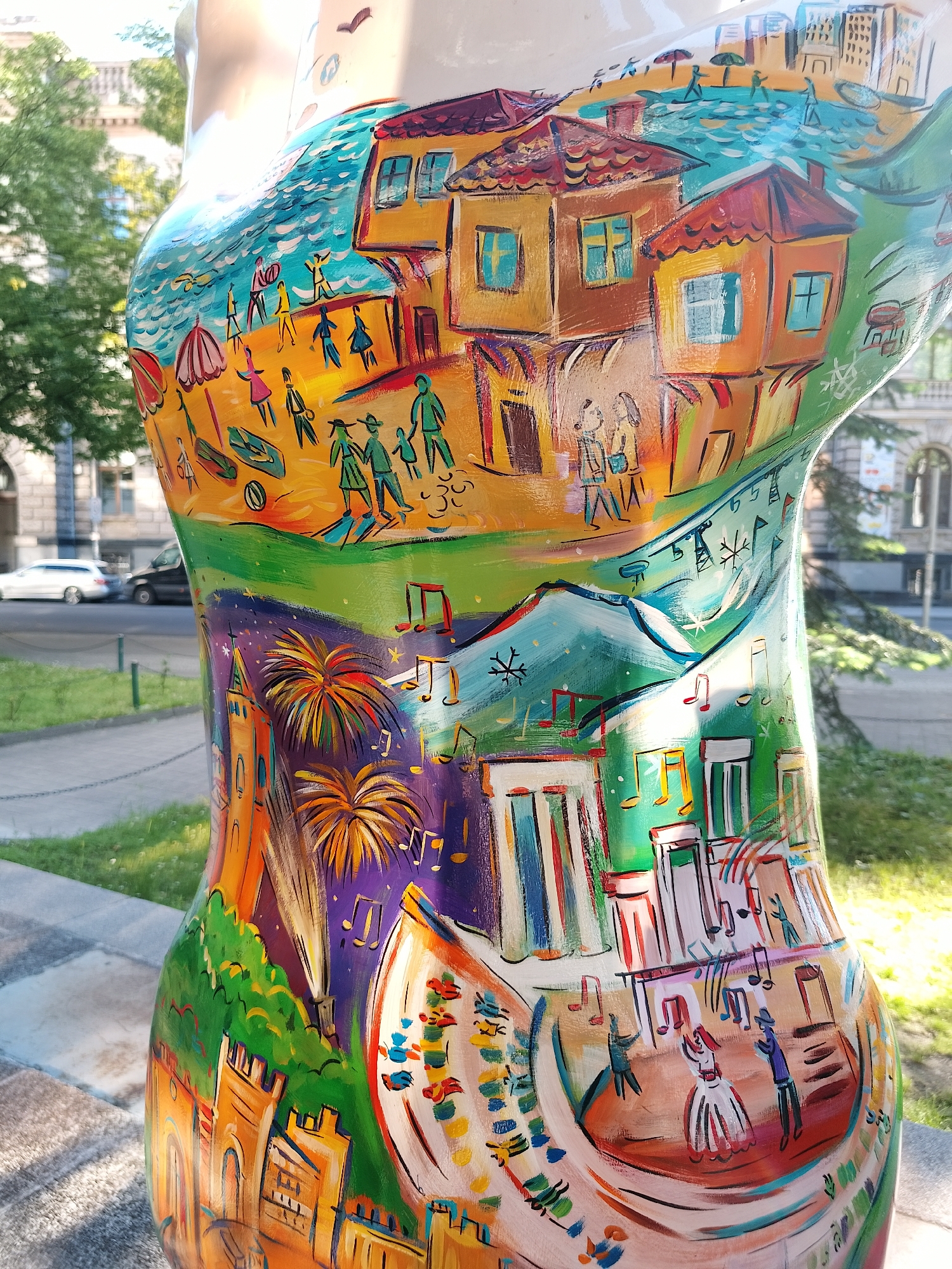 The image shows a torso sculpture with a colorful painting depicting various scenes, likely representing a city or region. The painting is divided into distinct layers or sections. The top section shows a beach scene with people, umbrellas, and boats. Above it, there is a depiction of traditional houses with red tile roofs. Below the beach scene, there is a vibrant layer with music notes and a cityscape with fireworks. The bottom section features a wedding scene with dancing figures and a castle-like structure. The sculpture is outdoors, likely in a park, as there is grass, trees, and a sidewalk in the background. In the distance, there are buildings and cars, suggesting an urban environment. The sculpture is likely part of a public art installation, showcasing different aspects of the city or region.