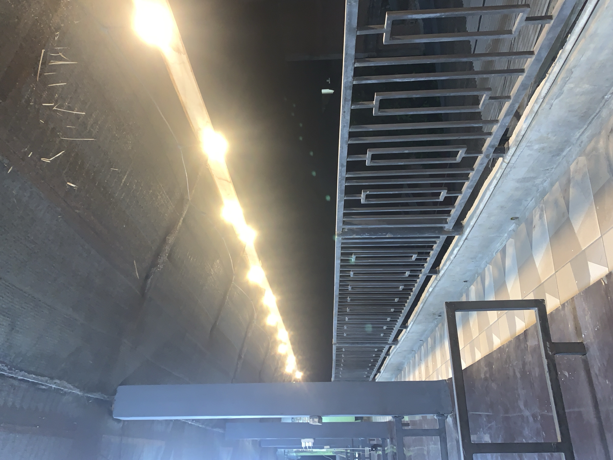 This image depicts an outdoor walkway or balcony area under a dark, textured ceiling, possibly a roof or awning. A line of bright, warm lights is strung along the underside of the ceiling, casting a glow. A dark metal railing with a geometric pattern runs along the edge of the walkway. The walkway itself appears to have a tiled floor with a diamond pattern in shades of beige and white, catching the light and casting shadows. To the left, a large, dark grey rectangular pillar supports the ceiling structure, and beyond it, parts of tables or seating are visible, appearing to be made of dark wood or metal. The background is dark, suggesting it is nighttime or a dimly lit area, with some faint green or white lights visible in the distance. The scene appears to be part of a public or commercial space, possibly a restaurant, bar, or entertainment venue in Palohlada, Indonesia. There are no people visible in the image.
