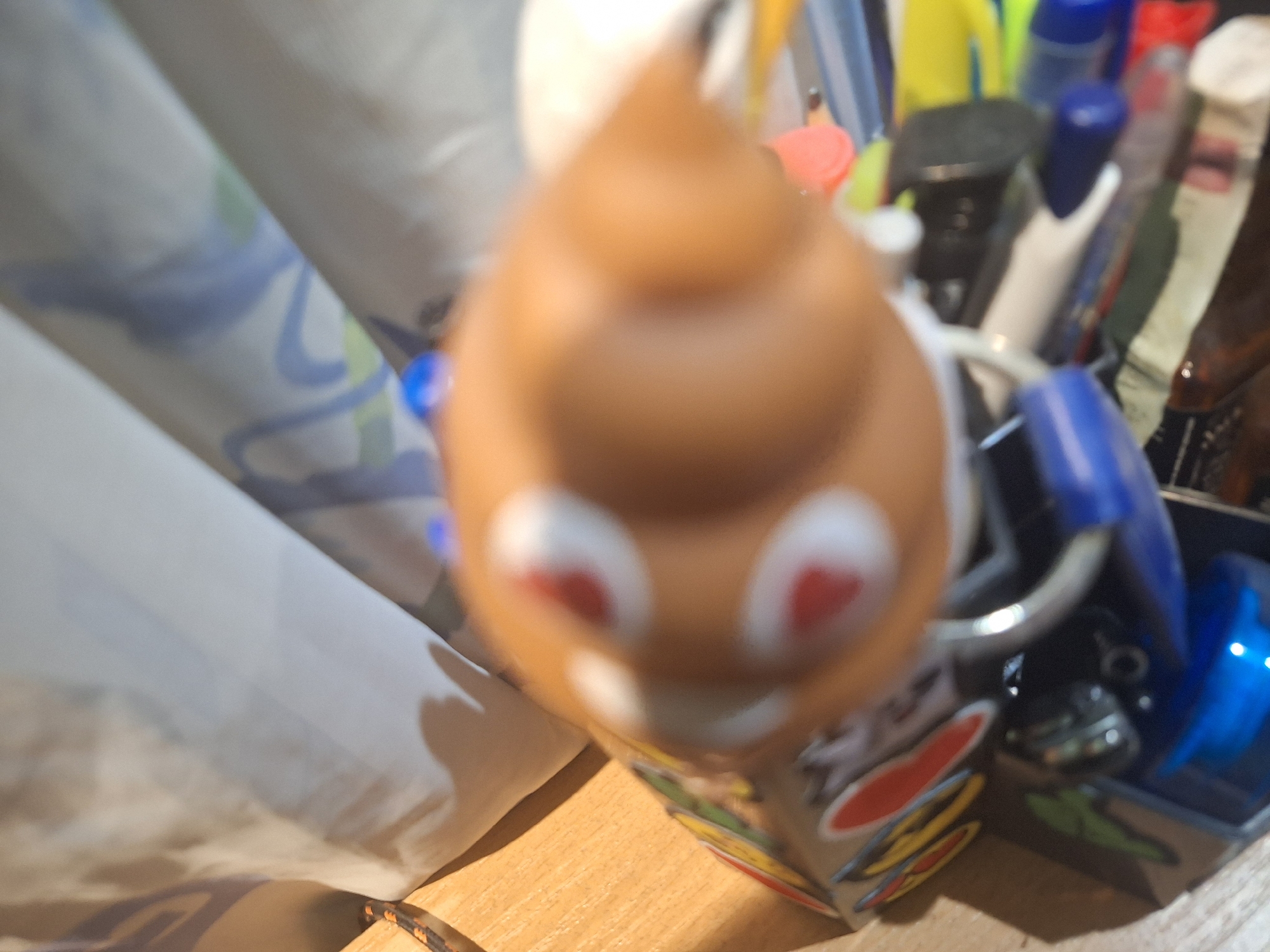 The image shows a close-up, blurry view of a light brown, rounded object with two red and white circular markings that resemble eyes. The object is positioned above a collection of pens and markers of various colors, including blue, yellow, and green, some of which have visible caps. The items are resting on a light-colored, textured surface, possibly a desk or shelf. In the background, a creased, light-colored fabric with blue and green patterns is visible. The lighting suggests an indoor setting, with a shadow cast on the left side of the frame. There is no indication of the city of Yambol, Bulgaria, or any other specific geographical location within the image itself.