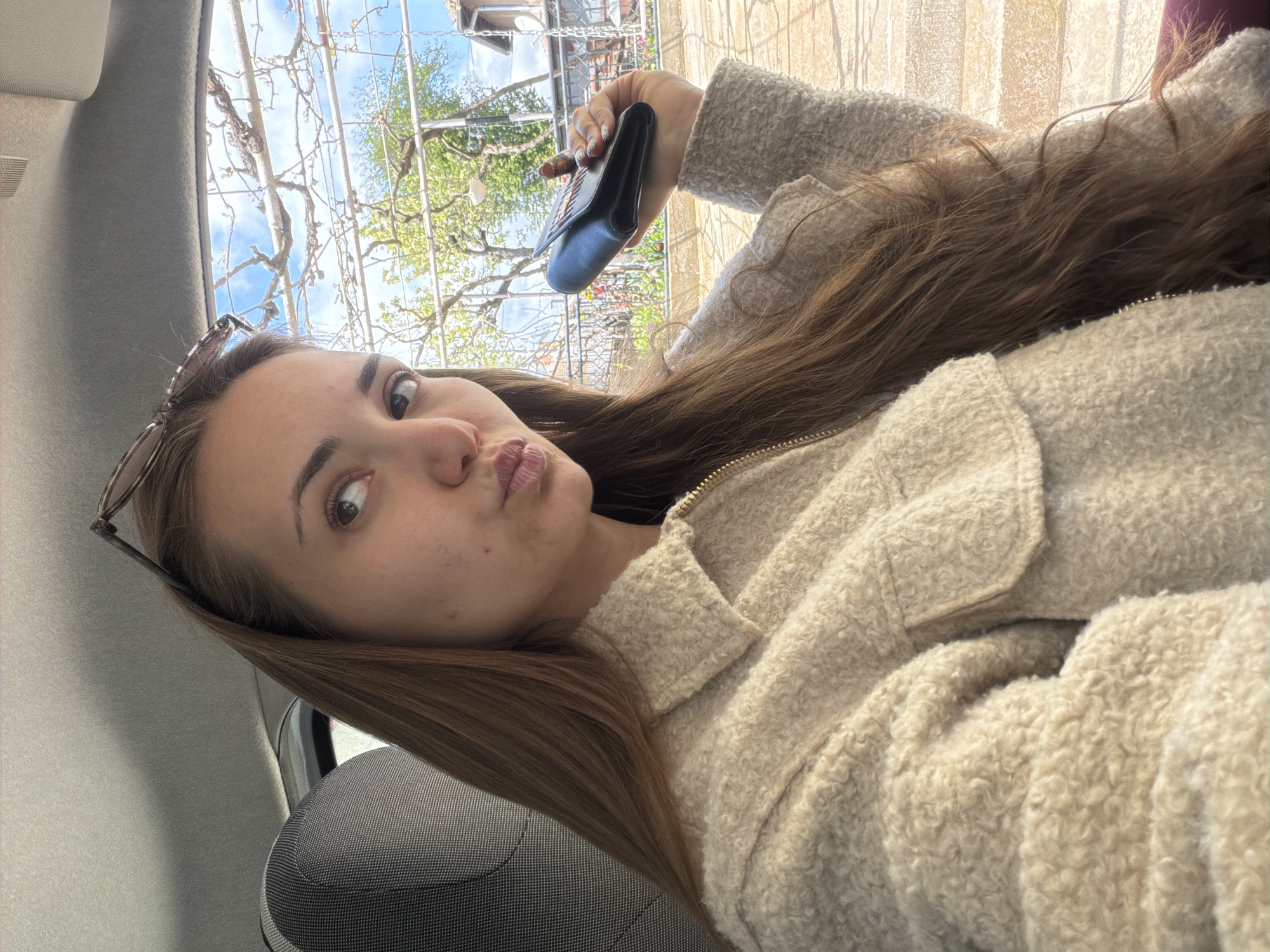 A young woman is lying down, likely in a car, holding a black wallet. She is wearing a light-colored, textured jacket and has brown hair. A pair of glasses are perched on her head. The view outside the car window includes trees and a structure with chains, possibly in Buhovo, Bulgaria.