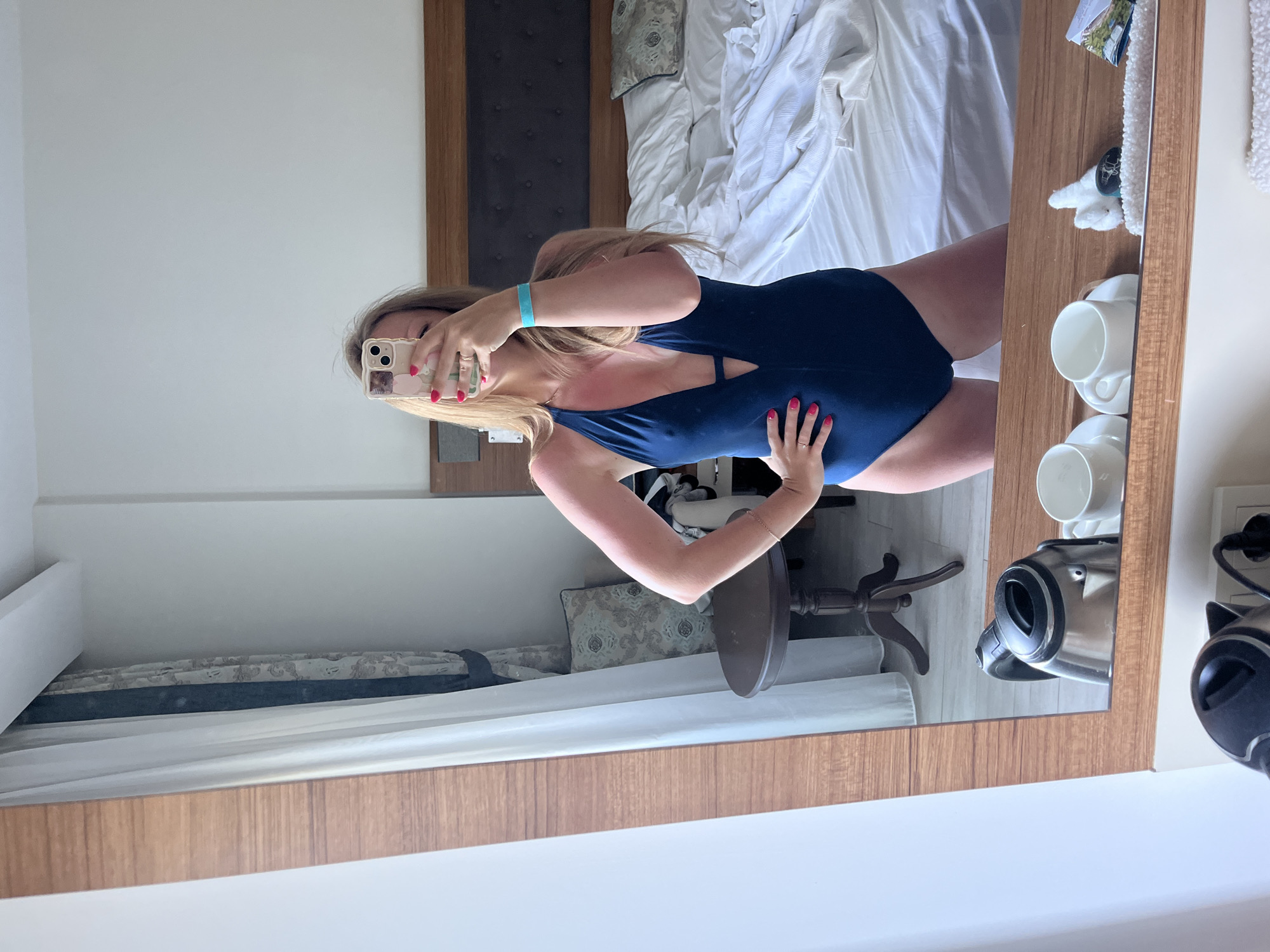 A blonde woman wearing a navy blue one-piece swimsuit and a turquoise wristband takes a mirror selfie in what appears to be a hotel room in Alanya, Turkey. She has bright pink fingernail polish and is wearing a ring on her left hand. Her left arm is on her hip, and her right arm is extended, holding up a phone to take the picture. Redness from sunburn is visible on her upper chest and shoulders. In the reflection, a bed with white linens is visible, along with a dark wooden headboard, a decorative pillow, a small round side table, and curtains. In the foreground of the mirror's reflection, a silver electric kettle and two white cups are on a wooden shelf. The room has light-colored walls and wood-paneled trim around the mirror.