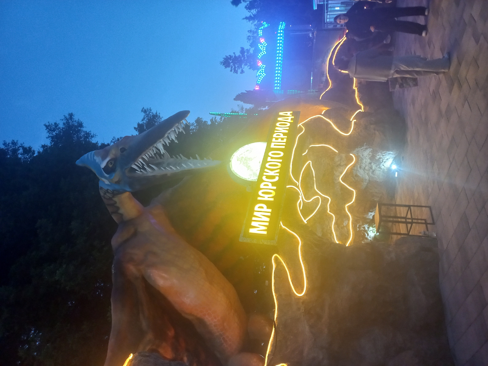 This image captures an evening scene at a dinosaur-themed attraction, likely in an amusement park in Sochi, Russia.
**Subjects:** The central subject is a large, light-blue statue of a pterosaur-like dinosaur with an open mouth revealing sharp teeth, positioned on the left side of the frame. In the background, several dark trees are visible against the twilight sky. On the right, three people are present: one woman in a dark jacket and pants stands observing the display, while another individual in lighter clothing walks away on a paved path. A third person is partially obscured behind them.
**Objects:** A large, bright yellow rectangular sign glows prominently with Russian text. Adjacent to this, yellow neon-like lights outline shapes on a dark, rocky-textured wall. Faint blue and multi-colored string lights are visible in the distance, suggesting other illuminated park features. The ground where the people walk is paved with a distinctive cobblestone pattern.
**Scene/Setting:** The scene is set outdoors at what appears to be a themed amusement park, specifically a "Jurassic Period" or "Jurassic World" area, as indicated by the signage and dinosaur statue. The sky is a deep blue, marking the transition from twilight to night.
**Activity or Event:** People are seen walking through and observing the illuminated dinosaur display, suggesting a leisurely recreational activity in an amusement park setting.
**Notable Details:**
* **Time of day:** Evening or early night, characterized by the dark blue sky and reliance on artificial lighting for visibility and atmosphere.
* **Weather:** The sky is clear, with no signs of precipitation.
* **Location cues:** The visible text in Russian on the sign and the provided context indicate the location as Sochi, Russia.
* **Emotions:** The people appear to be casually enjoying the attraction.
**Visible Text:** The bright yellow sign clearly displays the Russian text: "МИР ЮРСКОГО ПЕРИОДА," which translates to "WORLD OF THE JURASSIC PERIOD" or "JURASSIC WORLD."