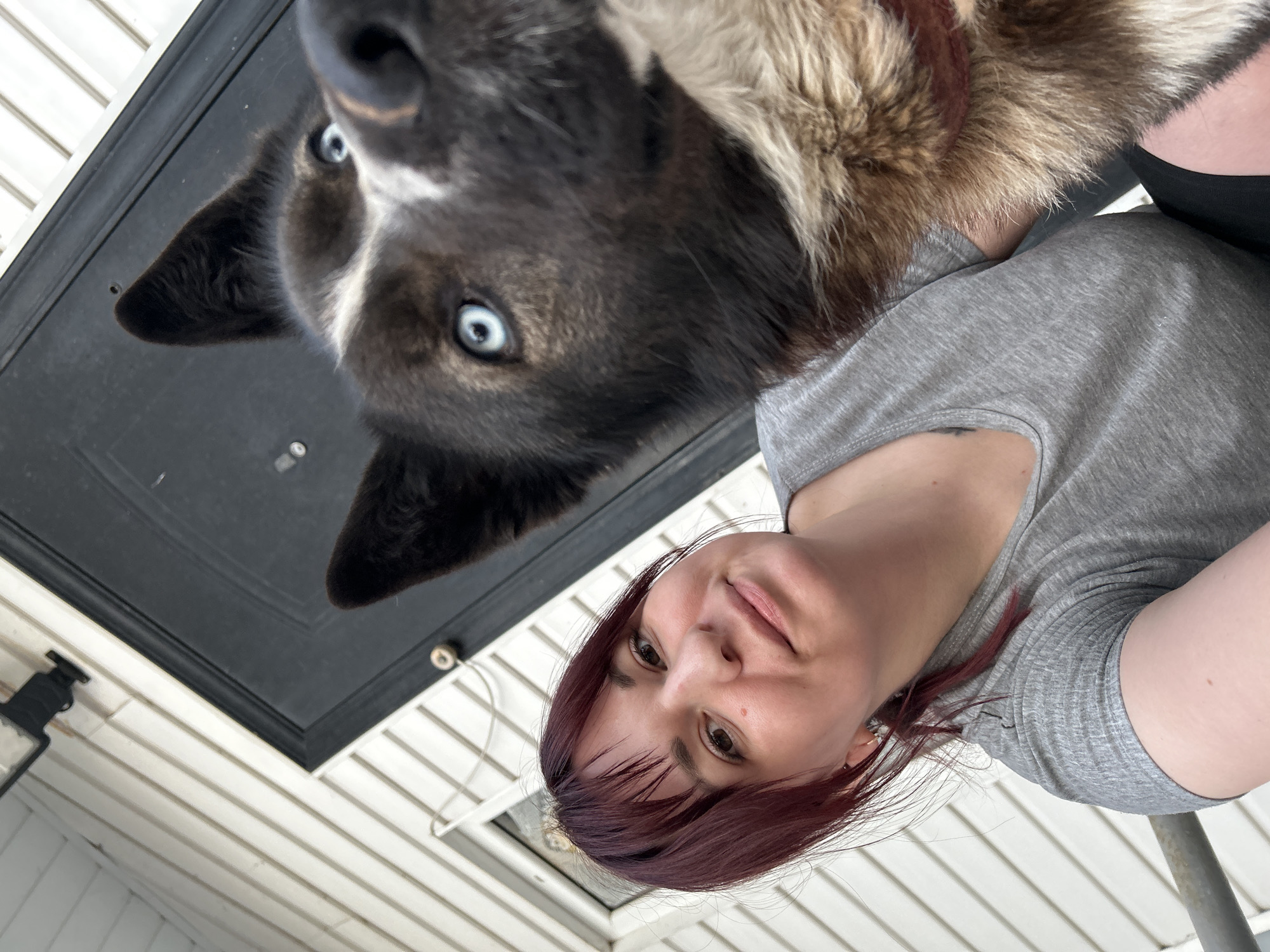 A woman with reddish-brown hair and a gray t-shirt is taking a selfie from a low angle, partially showing her shoulder and arm. A dog with blue eyes and black and tan fur is positioned above and slightly behind her, with its face in the upper left of the frame. A black door with a molding frame is visible behind the dog. White horizontal siding is visible in the lower half of the image, indicating an exterior location. The scene appears to be outdoors. No indication of the location in the city of Фоки, Russia, is visible.
