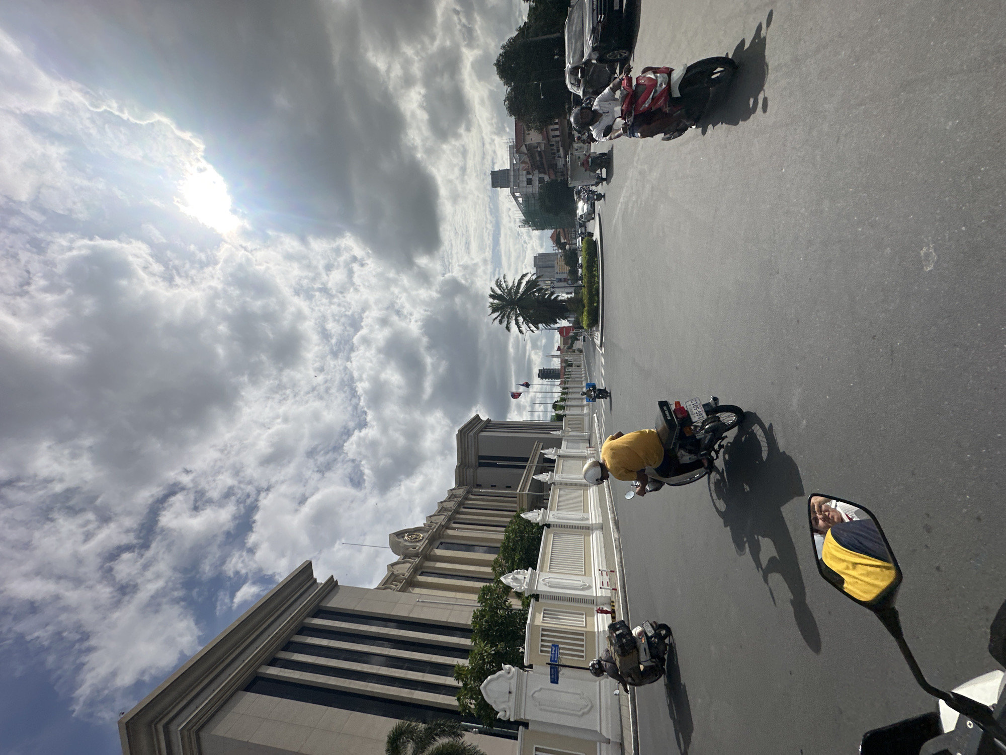 The image depicts a street scene in the Khan Daun Penh district of Cambodia. The focus is on a road with several people riding motorcycles. In the foreground, a rider in a yellow shirt is on a motorcycle, with their reflection visible in the side mirror. Further down the road, other individuals on motorcycles are also present, navigating the street.
To the left of the road, there is a large, ornate building with a light-colored facade and dark vertical accents. It is bordered by a white fence with decorative pillars. Trees are visible behind the fence, providing some greenery.
The sky is filled with dramatic clouds, with sunlight breaking through in the upper right portion of the frame, creating lens flare effects. In the background, other buildings and palm trees can be seen, suggesting an urban environment. The road surface is asphalt, and the overall impression is of a busy street on a cloudy but bright day.