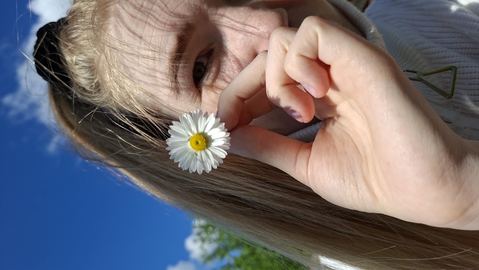A close-up, tilted portrait captures a young person outdoors under a bright blue sky with scattered white clouds. The subject, likely a child or teenager, has long, light blonde hair that appears highlighted by direct sunlight. Only part of her face, including her left eye, is visible, with some features obscured by shadows. She holds a small, white daisy with a vibrant yellow center between the index finger and thumb of her right hand, which is prominent in the foreground.
A black hair tie is visible in her hair near her ear. She is wearing a light-colored, possibly white, textured garment, which could be a sweater or blanket. A small, metallic, triangular-shaped object, possibly part of an accessory or clothing, is visible near her right hand resting on the white garment. The bright sunlight casts distinct shadows on her skin and hair, emphasizing individual strands. In the background, green foliage, likely trees, is visible beneath the sky, indicating a natural, open-air setting. The scene suggests a peaceful moment appreciating nature on a sunny day. No text is visible in the image. The location is Большой Букор, Russia.