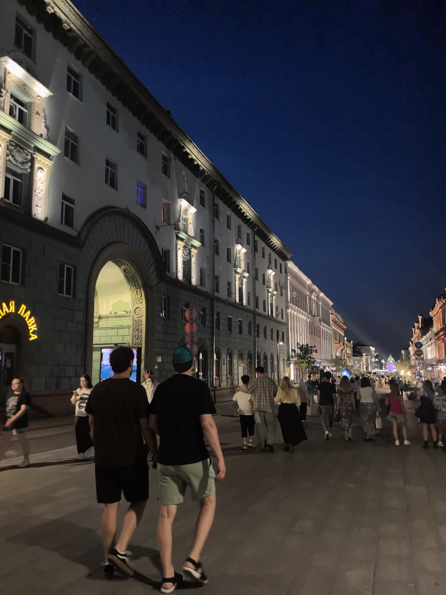 The image shows a street scene at dusk in Nizhny Novgorod, Russia, likely on Bolshaya Pokrovskaya Street, a popular pedestrian thoroughfare. A grand, multi-story building with arched entrances and ornate architectural details dominates the left side of the frame. Its windows are lit, casting a warm glow against the darkening blue sky. On the right, a bustling street unfolds, lined with buildings, shops, and a crowd of people.
In the foreground, two men walk away from the camera. The man on the left wears a black t-shirt and black shorts, with a black and white checked shirt draped over his left arm. He has black shoes with white soles. The man on the right wears a black t-shirt and light green shorts, with black sandals.
Further down the street, a diverse group of people are visible, many walking, some standing and conversing. They are dressed in casual summer attire. Shops with illuminated signs line the street, and streetlights cast a warm illumination on the scene. The overall atmosphere is lively and vibrant, suggesting a popular evening destination. The building on the left has a sign with yellow lettering that reads "ЛАЯ ЛАВКА" (Laya Lavka).