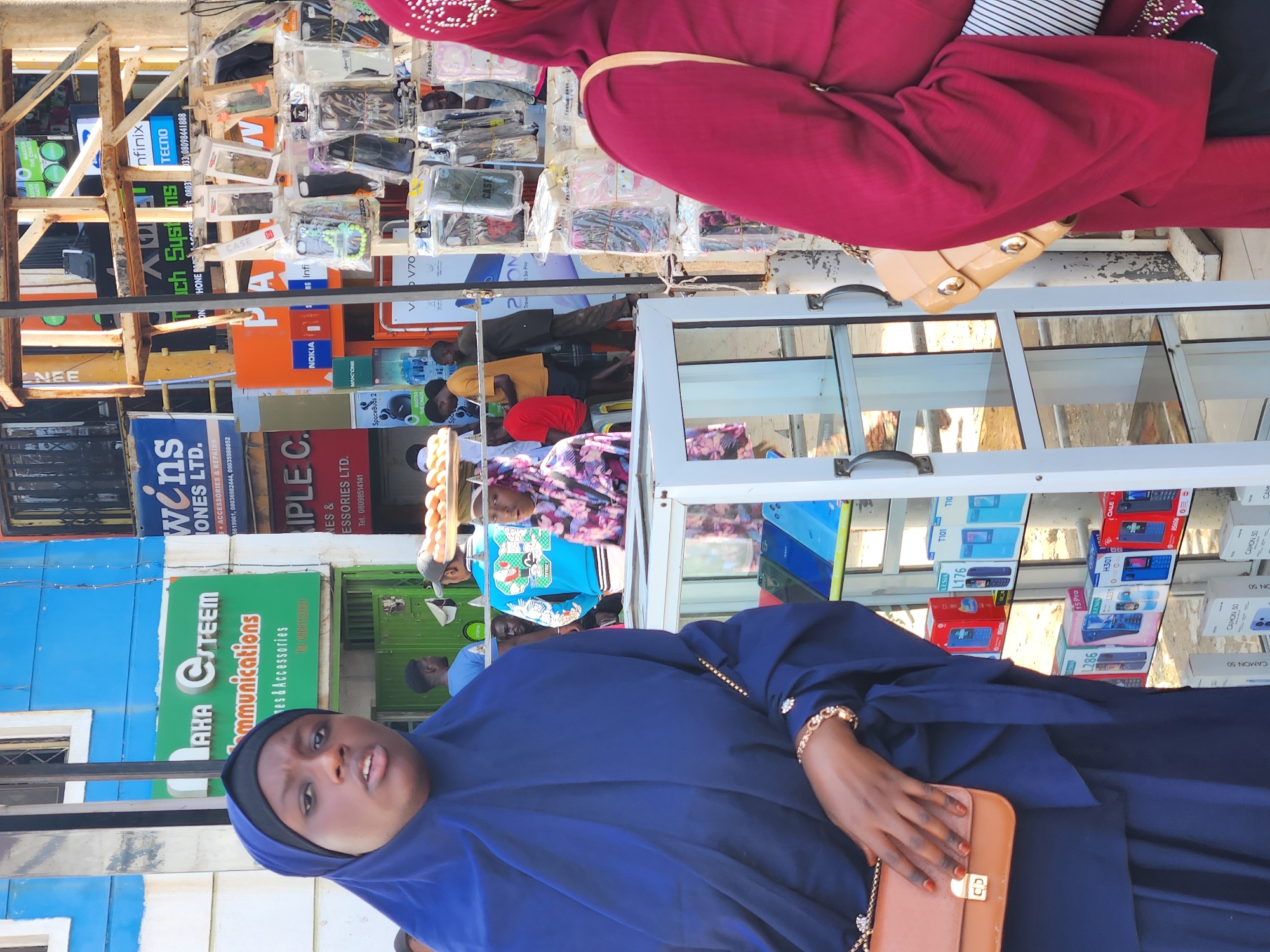 A medium-shot, eye-level outdoor photograph captures a street scene in Jos, Nigeria, focused on two veiled women and a stall displaying mobile phones and accessories.
In the foreground, a woman on the left wears a dark blue hijab and a matching abaya, carrying a tan handbag with a gold clasp. Her gaze is directed slightly off-camera to her left. To her right, a woman in a dark red hijab and abaya is partially visible, also with a tan handbag.
Behind them, a stall is laden with numerous phone cases packaged in clear plastic, hung on a metal rack. Below this, a glass display cabinet showcases several mobile phones of various brands and colors, arranged on shelves. In the background, a variety of signs are visible, including one with green lettering that reads "LAHA GSTEEM Communications" and another advertising "IPLE C." Other signs appear to promote mobile phone brands like Nokia and Infinix.
Several individuals are interspersed throughout the background, some appearing to be customers and others possibly vendors. One person in the mid-background is seen carrying a tray of eggs on their head. The overall environment suggests a busy marketplace or commercial area. The lighting indicates it is daytime.
