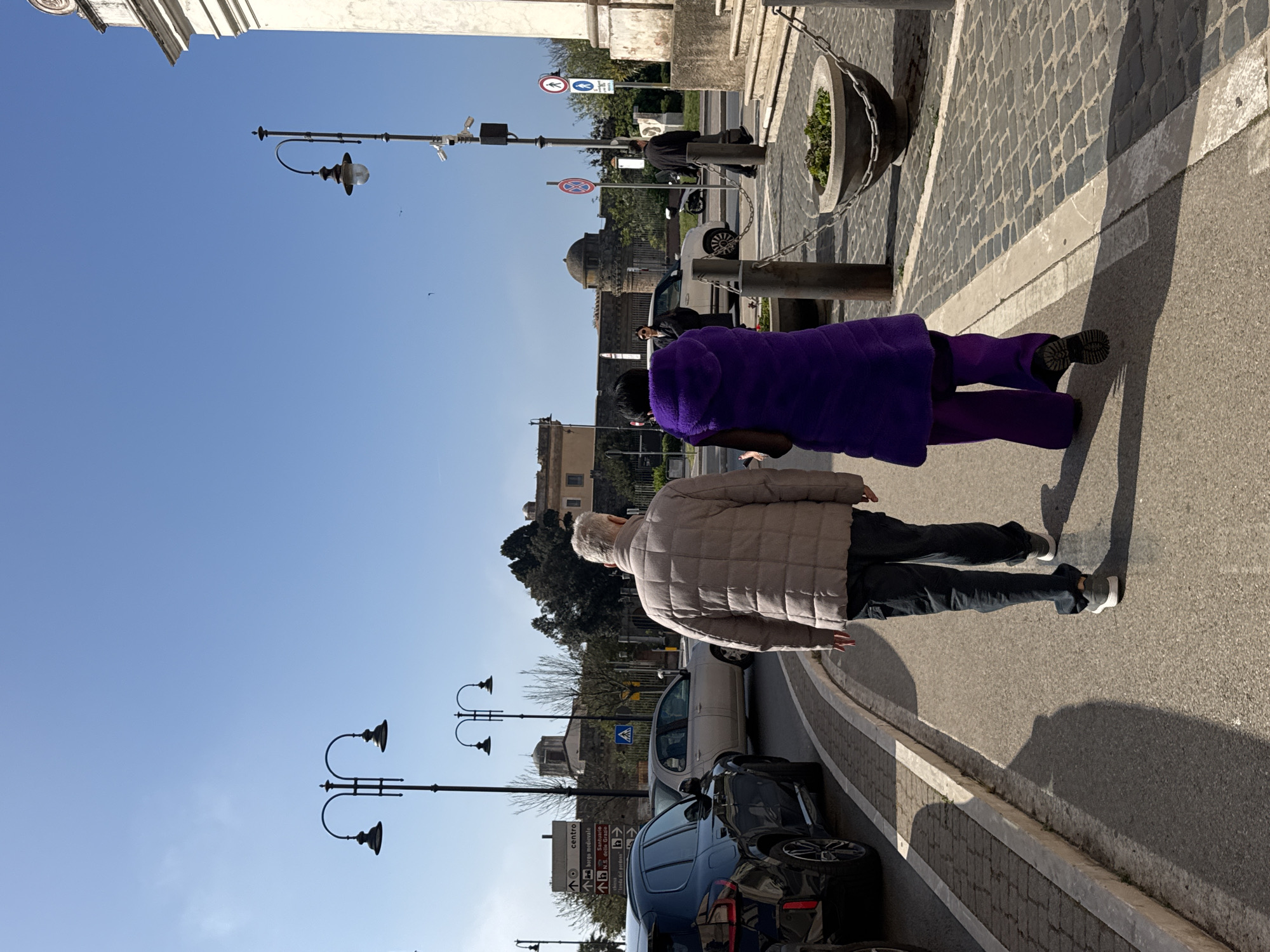 The image captures a street scene in Nettuno, Italy, on a sunny day. Two individuals are walking along the sidewalk adjacent to a road with parked cars. The first person is wearing a tan quilted jacket and dark pants and appears to be walking alongside the road. Behind them is a second individual wearing a purple fleece outfit. The road is lined with lampposts, and street signs are visible in the background. Across the road, there is a building with a dome. The sidewalk is paved with cobblestones.