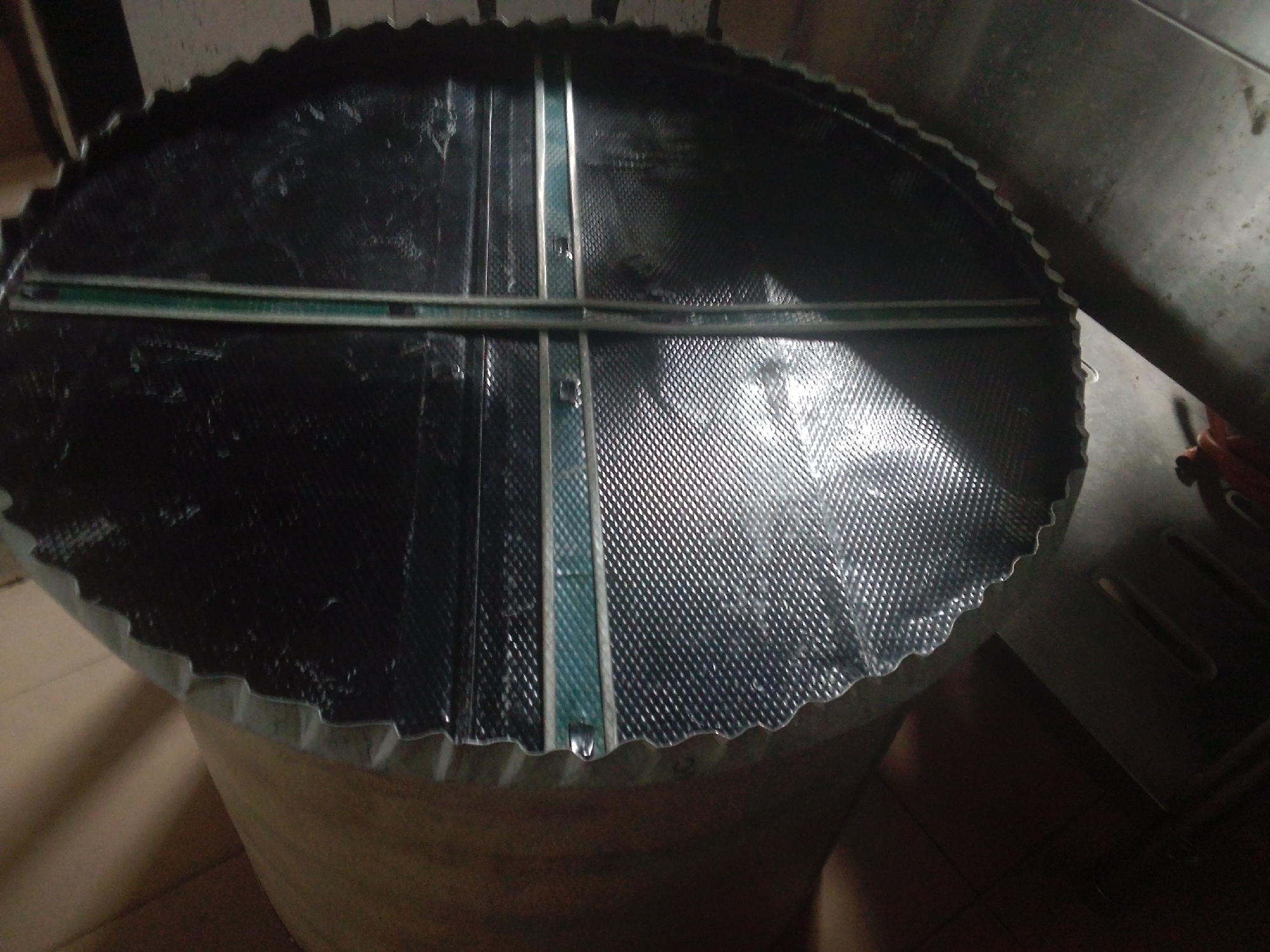 This media file shows a close-up, overhead view of the top of a large, cylindrical object, possibly a storage tank or industrial drum. The lid of the object is made of a dark, textured material, possibly plastic or a reflective metallic foil, with a pattern of small diamond shapes. Two metallic or plastic strips, one horizontal and one vertical, cross over the lid, dividing it into four sections. These strips appear to be secured with small fasteners or rivets. The edge of the lid is wavy or corrugated, with a crimped metal trim that appears to be attached to a lighter-colored, possibly cardboard or composite material, cylindrical body of the object.
The object is resting on a floor that appears to be tiled or made of smooth concrete. The lighting suggests an indoor setting, possibly a workshop or storage area. The overall impression is of a functional, industrial-style object, possibly in a state of assembly, repair, or storage. There is no visible text in the image. The lighting casts shadows, indicating it is likely daytime, though the specific time is not discernible. The scene does not contain any people. The location context is provided as Howayi, Nigeria, which could suggest the type of industrial or agricultural equipment found in that region.