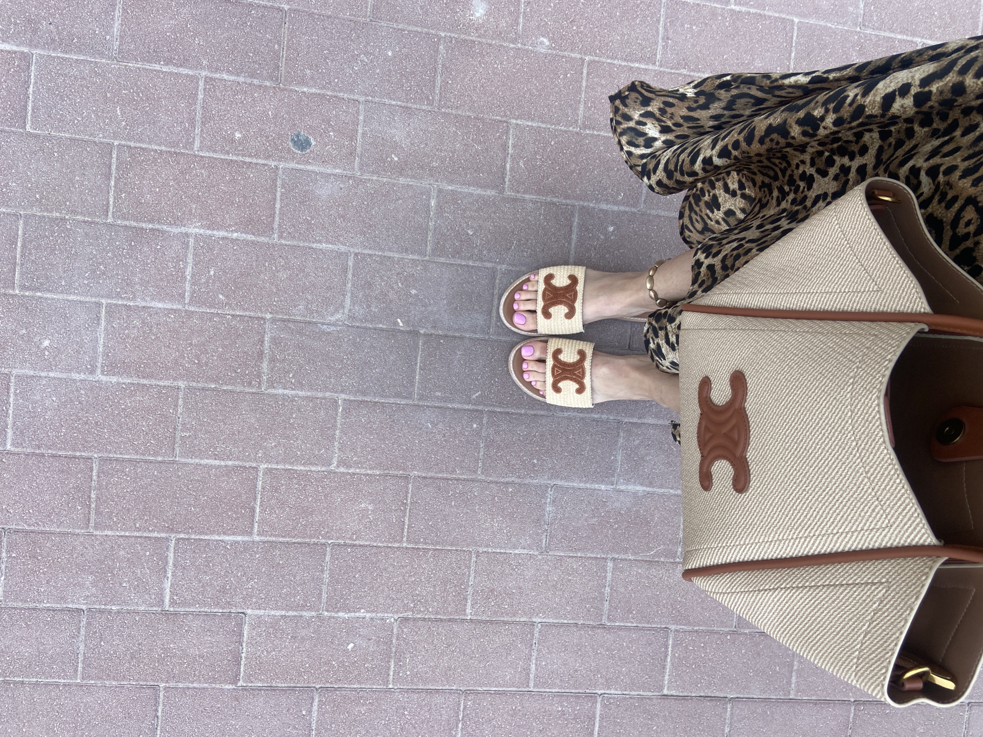 A high-angle view captures a portion of a person standing on a brick-paved surface, likely in Dubai, United Arab Emirates, indicated by the warm tones and modern urban design. The person wears an ankle bracelet and slides with a designer logo. The toenails are painted light pink. A leopard-print dress or skirt is visible, along with a large tote bag made of woven material with a leather designer logo. The tote bag reveals a glimpse of its interior, and is situated close to the person. The bricks on the ground take up a large portion of the frame, exhibiting a consistent pattern with visible grout lines. The overall lighting is bright, and the composition is dominated by the brick-paved surface, with the subject appearing on the right side of the frame.