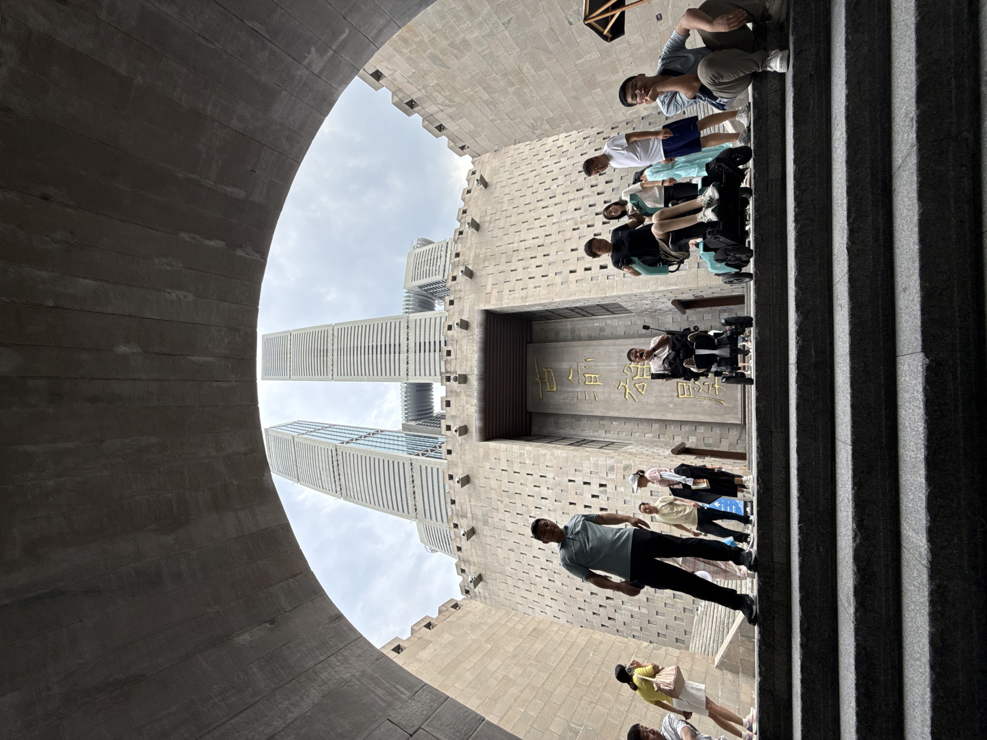 In the Nan'an District of China, a group of people are gathered on a wide, stone staircase leading up to a monumental stone structure. The structure resembles a fortified entrance with crenellations and a large, central doorway. Above this structure, two modern skyscrapers with glass facades rise into a cloudy sky.
Several individuals are positioned on the stairs and at the entrance. On the left, a person in a light blue polo shirt and black pants stands facing forward. Further up the stairs, to the right of the central entrance, a woman in a black shirt and shorts is seated in a teal electric wheelchair, with a person in a white shirt and a woman in a light blue garment standing nearby. To the far right, a young man in a light blue t-shirt and brown pants is sitting on the steps, resting his chin on his hand. Other people are partially visible further up the stairs and to the left.
The foreground is dominated by the dark, textured stone steps, suggesting a grand and imposing architectural design. The overall scene captures a moment of public gathering in a location that blends historical architecture with contemporary urban development.
