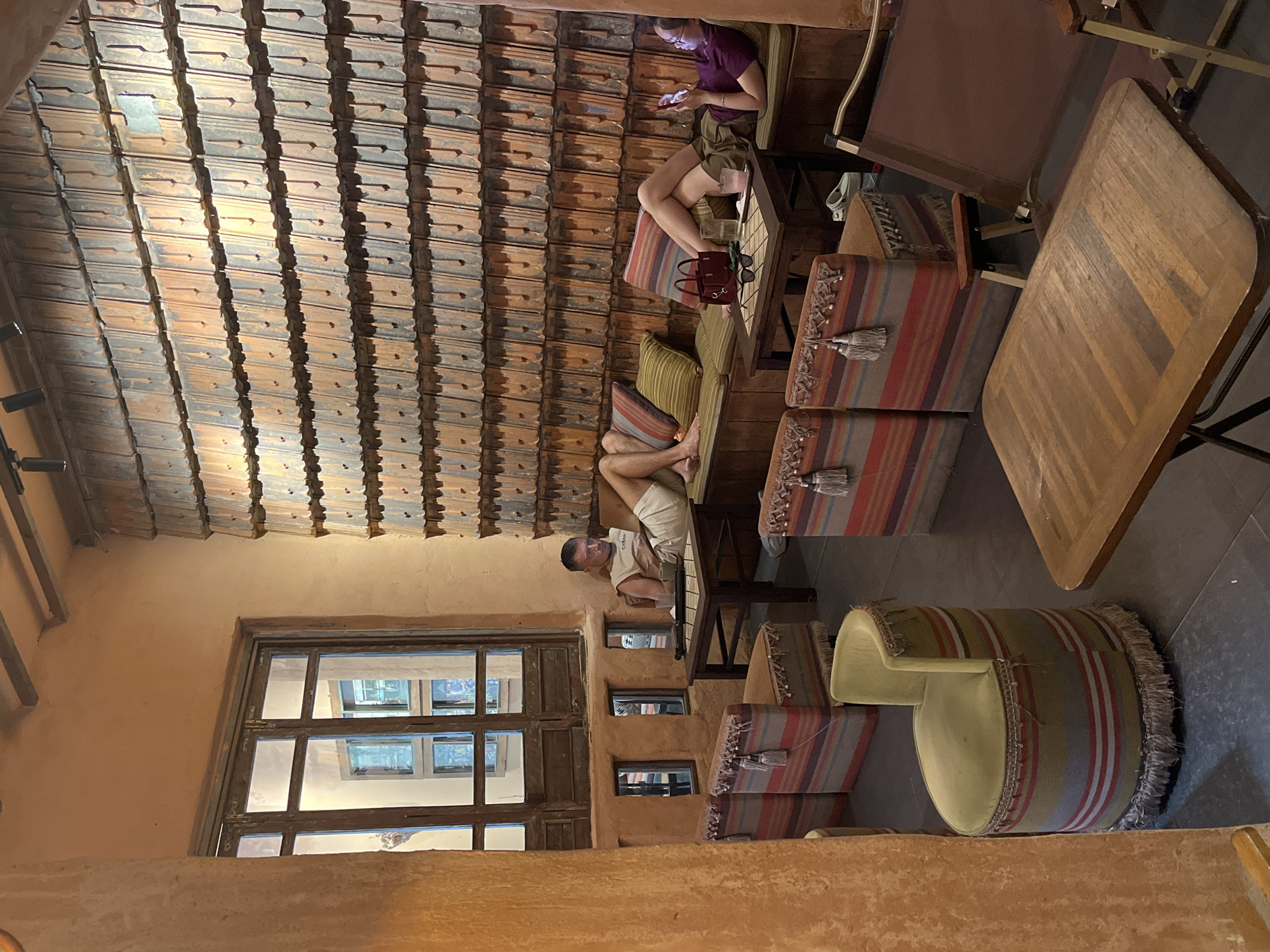 The image depicts the interior of a cafe or restaurant in Đà Nẵng, Vietnam, judging by the distinctive architecture and interior design. The restaurant features a low-ceiling roof constructed of terracotta tiles and tan-colored plastered walls. A large window with divided panes is visible, suggesting an exterior view.
Two people are present in the image. One person is sitting on a low cushioned bench while using a smartphone. Another person is lying down on a similar cushioned bench. Striped cushions and furniture are seen throughout.
Tables and chairs are interspersed with the cushioned seating areas. The furniture features a combination of wood and striped fabric. Overall, the scene appears to be a relaxed, informal setting.