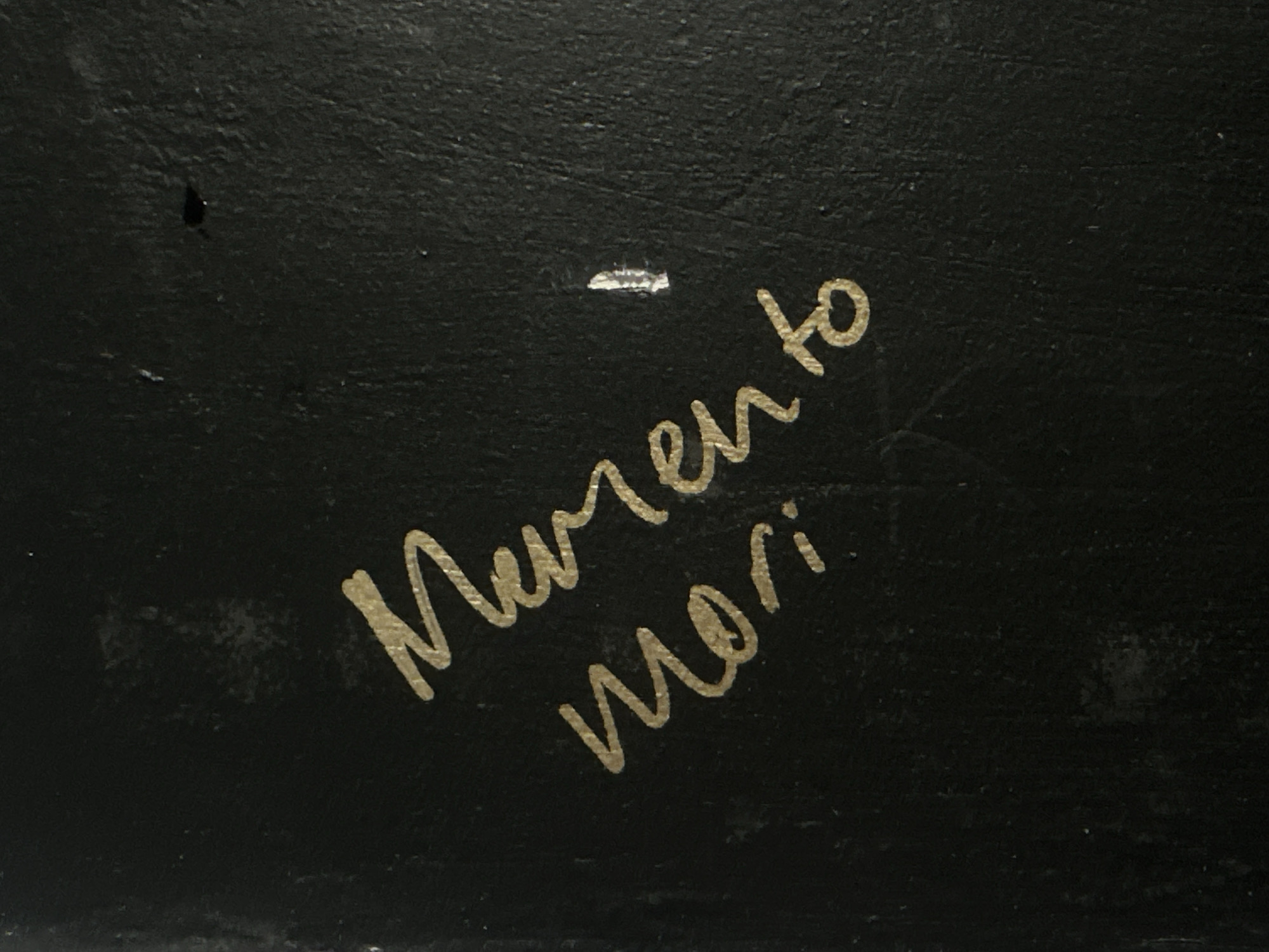 The image features light-colored, handwritten text against a dark, textured background. The background surface is black or very dark grey, exhibiting a slightly uneven or rough texture. Minor imperfections are visible on this surface, including a small dark spot in the upper left portion and a horizontal, light-colored mark situated above the text.
The visible text is written in a flowing, cursive script and reads "Memento Mori." The word "Memento" is on the upper line, while "mori" is on the lower line, positioned slightly to the right and below the first word. The text is rendered in a metallic gold or light yellow color, providing a strong visual contrast against the dark background.
No people or specific activities are depicted. The time of day or weather conditions cannot be determined from the image. While the location context is given as London, United Kingdom, the image itself does not contain any specific visual cues to indicate this location. The phrase "Memento Mori" is a Latin aphorism serving as a reminder of mortality.