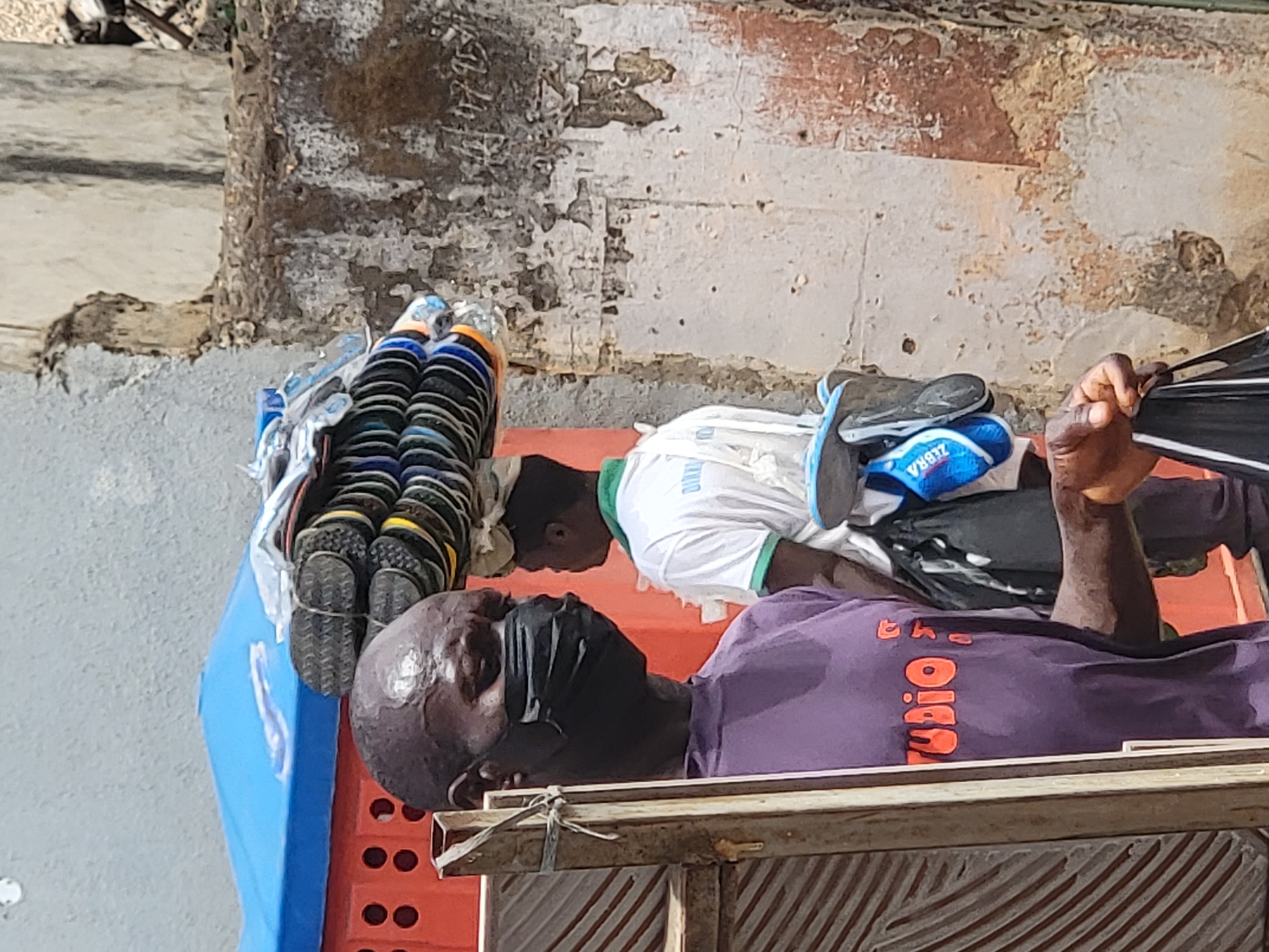 A vendor wearing a purple shirt and face mask lies on a wooden cot next to a rack of sandals for sale. Another individual in a white shirt with green trim is nearby. The setting appears to be an outdoor urban environment, possibly a street in Maiburiji, Nigeria. The wall in the background is distressed, with peeling paint and exposed concrete.