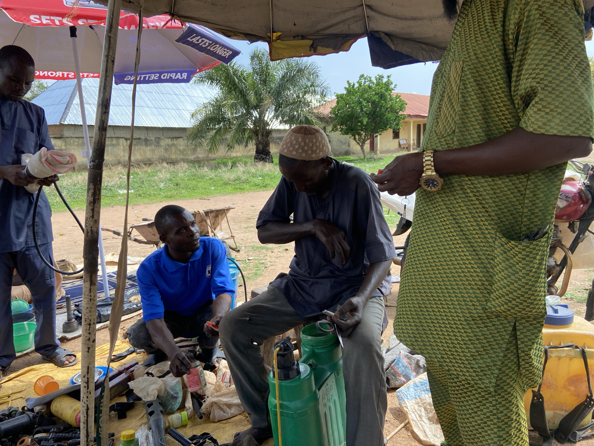 This image depicts an outdoor scene in Mararaba, Nigeria, likely a market stall or repair station, where four men are engaged in various activities under makeshift shelters. The setting is characterized by a mix of natural elements and simple man-made structures.
In the foreground, a man wearing a light-colored cap and dark clothing is seated, appearing to be mending or assembling something with small tools in his hands. Next to him is a green backpack-style sprayer. To his left, another man in a blue polo shirt crouches, observing and holding a small red object. On the far left, a man in a blue traditional outfit is standing, holding a bundle of white and red material, possibly wires or textiles. Partially visible on the right is a fourth man, standing, wearing a green and yellow checkered patterned attire, a prominent gold watch on his left wrist, and holding a small red item in his hand.
The ground around them is a bustling array of objects, including plastic containers, bags, tools, and various machine parts, indicating a commercial or repair-oriented environment. Overhead, two umbrellas provide shade from the sun; one red and white, displaying reversed text like "SET" and "STRON", and another blue and white with reversed text "LASTS LONGER" and "RAPID SELLING".
The background reveals a grassy area, scattered trees including palm trees, and modest buildings with corrugated iron roofs, indicative of a rural or semi-urban African landscape. The clear skies and strong shadows suggest it is daytime, likely mid-day. The men appear focused and engaged in their tasks, contributing to a sense of everyday commerce and work.