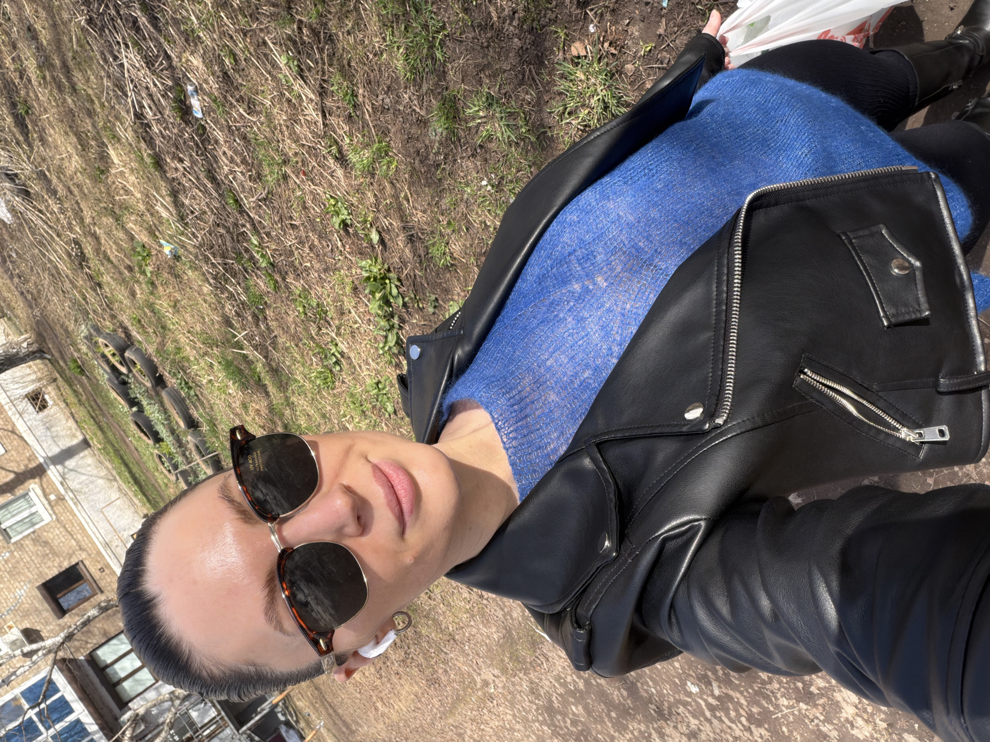 A woman wearing sunglasses, a blue sweater, and a black leather jacket poses for a selfie. She is wearing an earbud in her right ear. She is also wearing black pants or leggings and black boots. She is holding a white plastic bag.
The background consists of patchy grass and some dry foliage. A multi-story building is visible on the left side of the frame.
The location is Perm, Russia.