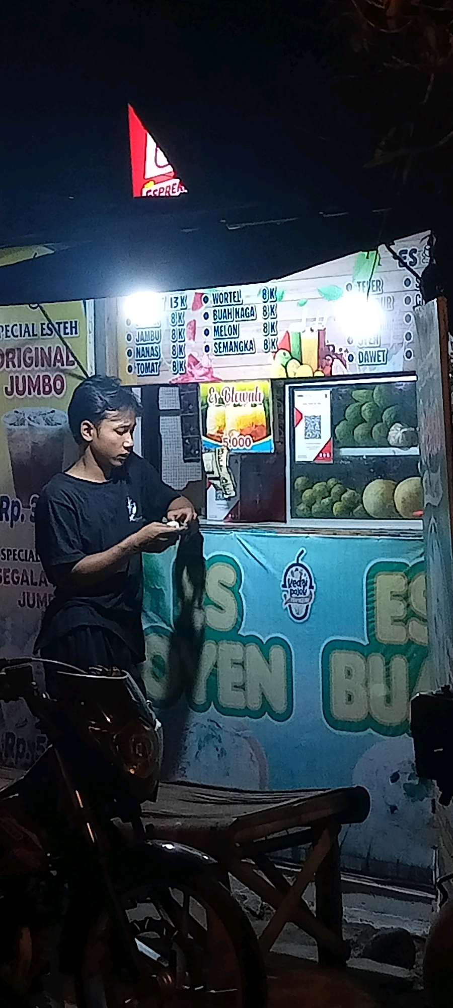 The image depicts a street-side food stall in Singogalih, Indonesia, at night. A young man, likely an employee, is standing behind a counter, holding what appears to be small coins and a small white object in his hands, possibly engaged in a transaction. The stall is illuminated by overhead lights, highlighting a menu board displaying various drink options and prices, including "Es Blawuh Rp 5.000". Various fruits like durian and avocado are visible in display cases. A motorcycle is parked in the foreground. The stall has a blue banner with large white lettering, and a smaller logo of a cup with the text "Kedai Pojok" is attached to it. The overall impression is a lively, local eatery operating in the evening.