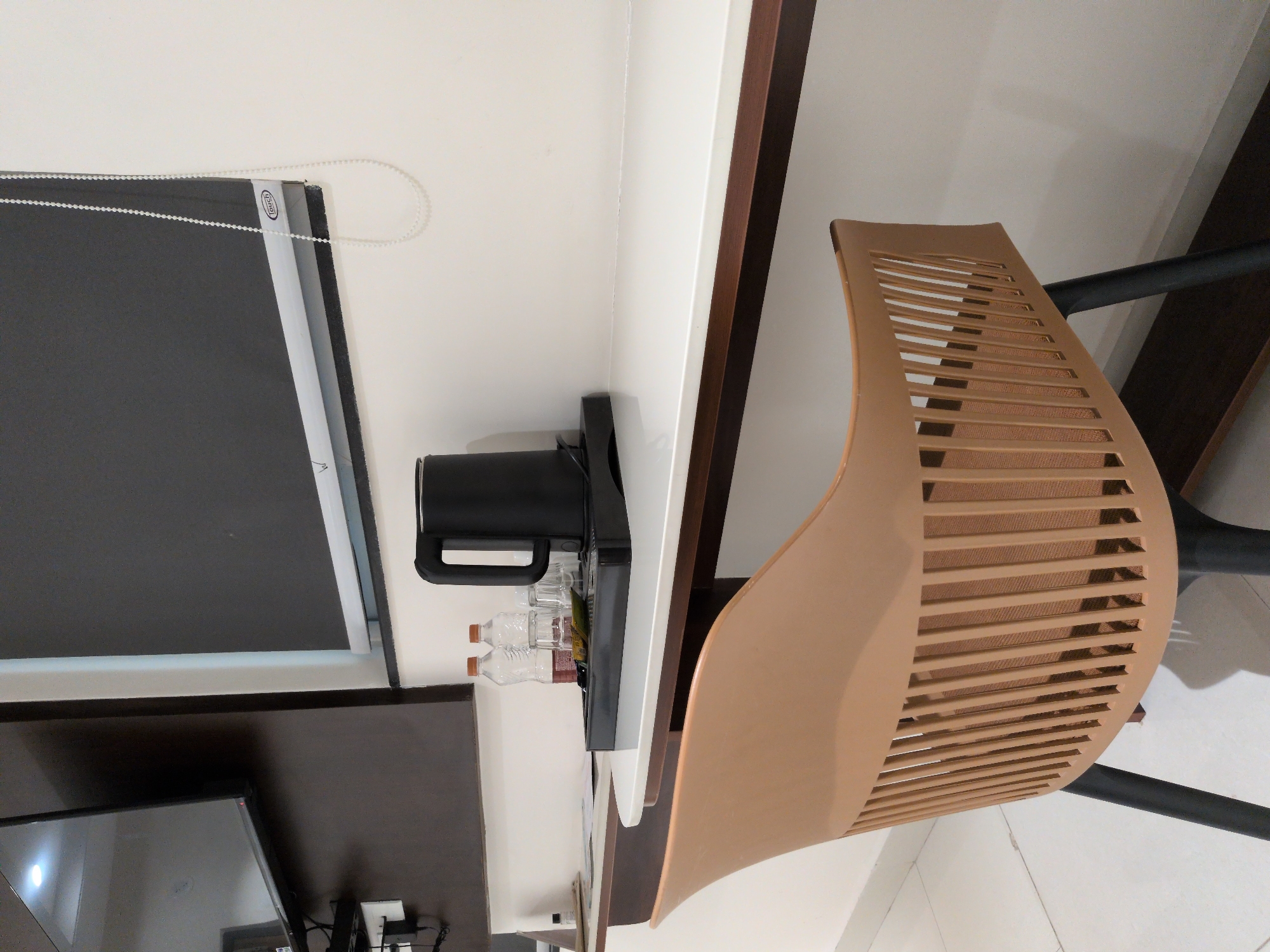 This indoor forensic image, captured in Hyderabad, India, depicts a static, unoccupied scene featuring a functional desk area. In the foreground, a light beige, modern-style chair with a slatted back and dark grey legs is positioned with its back to the viewer, facing a white-surfaced desk or counter. On the desk, a black tray holds a black electric kettle, two transparent plastic bottles with orange caps containing a reddish-brown liquid, and two clear drinking glasses. A small green sachet is also on the tray. To the left of the tray, several papers or brochures are visible. The wall behind the desk is plain white. To the upper left, a wall-mounted, dark-screened flat-panel television is partially visible, reflecting an internal light source. Below the television, a black shelf or bracket supports a power outlet or similar electrical fixture with associated cabling. In the upper center-right, a dark grey roller blind, with the brand name "Touch" visible on its lower edge, covers a window. A white beaded chain, used for operating the blind, hangs vertically on the right side. The flooring is a light-colored tile. No individuals are present in the frame.