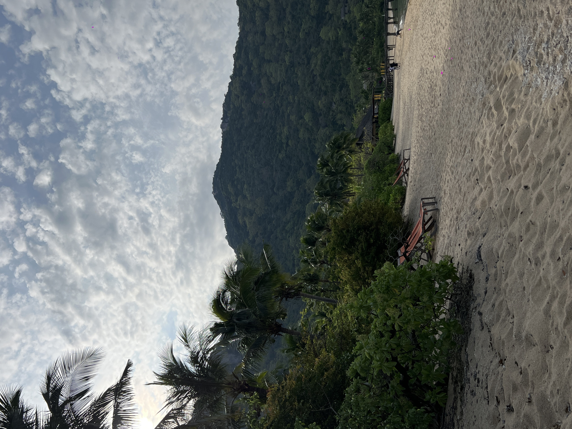 The image captures a tranquil beach scene in Đông Ninh Hòa, Vietnam. The foreground is dominated by soft, light-colored sand, showing gentle ripples and some scattered debris. Two empty orange lounge chairs are positioned on the sand, hinting at relaxation. Lush green vegetation, including palm trees and dense bushes, lines the edge of the beach, leading towards a forested mountain that rises in the background. The mountain is covered in a thick canopy of trees. A wooden pier extends into the water on the right side of the frame, where a few indistinct figures can be seen walking. A small thatched-roof building with yellow awnings is visible near the pier, suggesting a resort or dining area. The sky is partially cloudy, with textured white clouds against a pale blue backdrop. Sunlight filters through the clouds, casting a soft light on the scene.