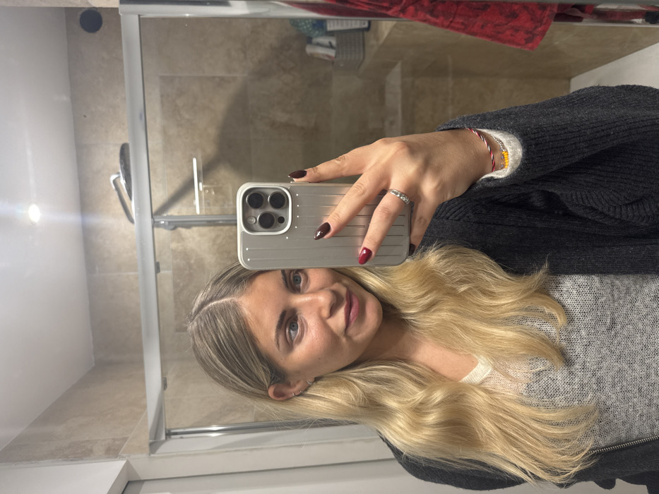 A woman with long blonde hair and fair skin takes a selfie in a bathroom mirror. She is wearing a gray sweater and a dark cardigan. Her nails are painted a deep red. She holds a silver phone with a triple-lens camera. A ring adorns her finger, and a colorful beaded bracelet is visible. The background features a shower stall and a beige-tiled wall.