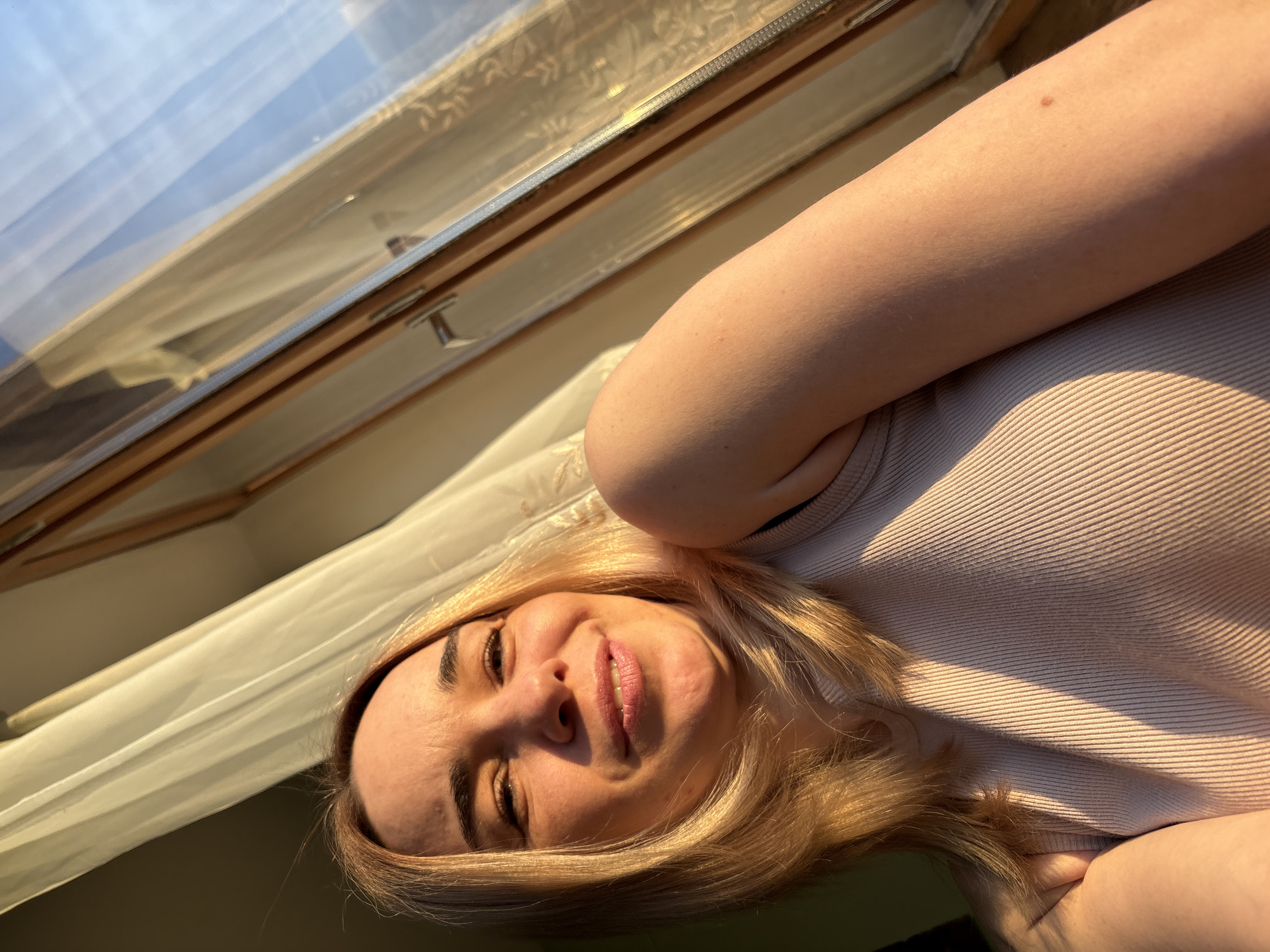 An indoor scene in Sinyavino, Russia, shows a woman bathed in the warm, golden light of either sunrise or sunset. The woman, who appears to be lying down or reclining, is the primary subject, with her head and upper torso occupying the lower right portion of the horizontally oriented image. She has light blonde hair and dark eyebrows, and a gentle, pleasant smile with slightly parted lips. Her skin is illuminated by strong, direct sunlight, which highlights facial features and casts subtle shadows. She is wearing a light-colored, ribbed top, across which distinct diagonal streaks of sunlight and shadow fall. A small, dark mole or freckle is visible on her upper arm.
In the upper left of the frame, a window, possibly a skylight, is prominent. It features a visible wooden frame and a metal latch or handle. Through the glass, a bright, clear sky is visible, along with reflections. Light-colored curtains or fabric are draped around the window, catching the golden light. The overall mood conveyed is one of relaxation and calm, with the natural light creating a soft, ethereal atmosphere. There is no visible text in the image.
