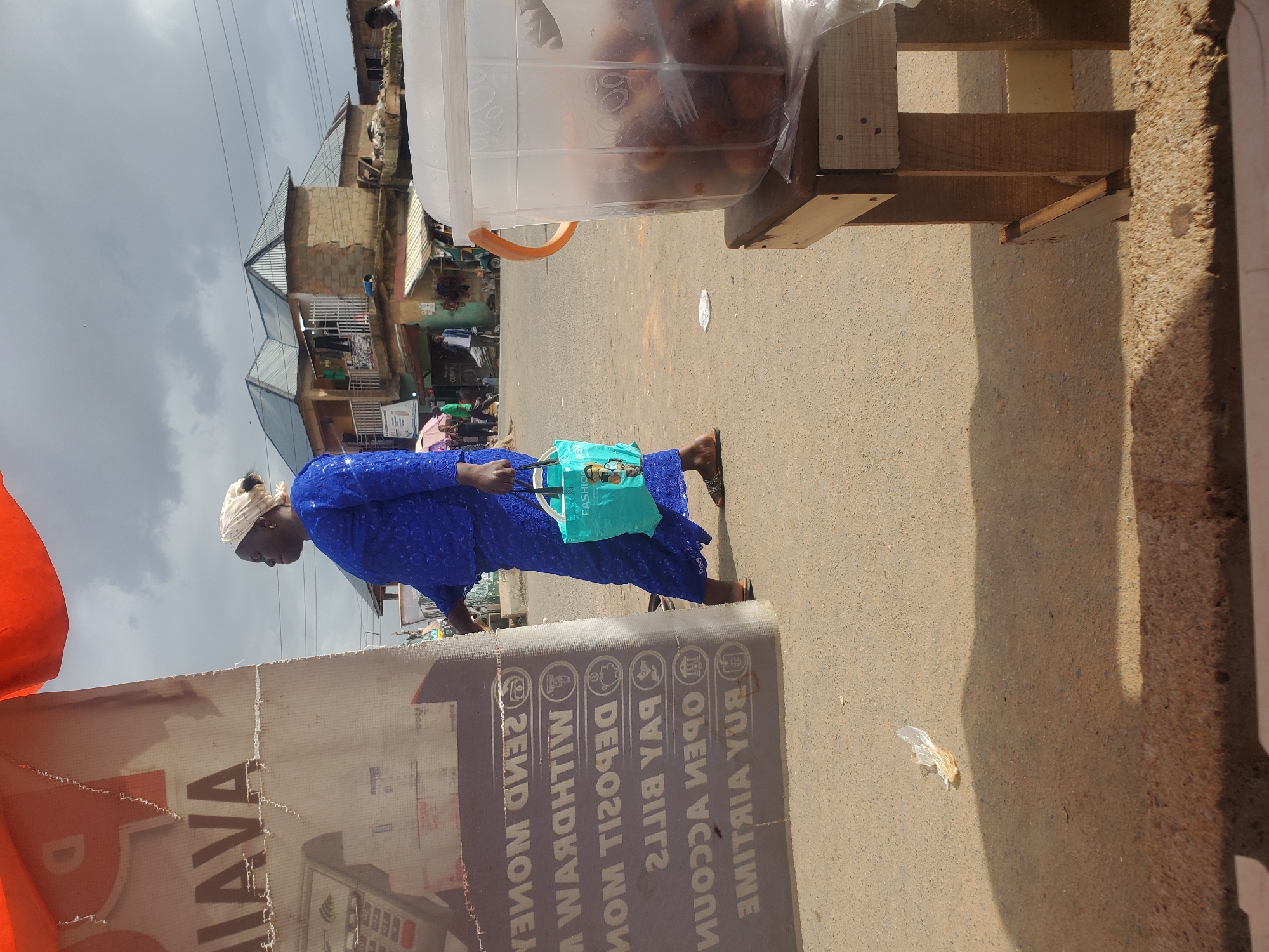 The image depicts an outdoor scene in Gwong, Nigeria. A woman, dressed in a blue traditional outfit and a light-colored headscarf, is walking on a paved surface. She is carrying a light blue bag. In the background, there are buildings with corrugated metal roofs, indicative of local architecture. The sky is overcast, suggesting a cloudy or potentially rainy day.
To the right of the woman, a wooden stand holds a large, clear plastic container filled with food items, possibly pastries or fried dough balls. The stand is partially visible, made of rough-hewn wood.
In the foreground, a large, faded banner is partially visible, with text that appears to be in a Western script, possibly English. Some of the legible text includes "JAVA", and further down, "BUY AIRTIME", "OPEN ACCOUNTS", "PAY BILLS", and "SEND MONEY". This suggests the banner is advertising some form of financial services or a business.
The overall scene appears to be a street or market area, with a mix of residential or commercial structures and everyday street life. The time of day is not explicitly clear due to the overcast conditions, but it seems to be daytime.