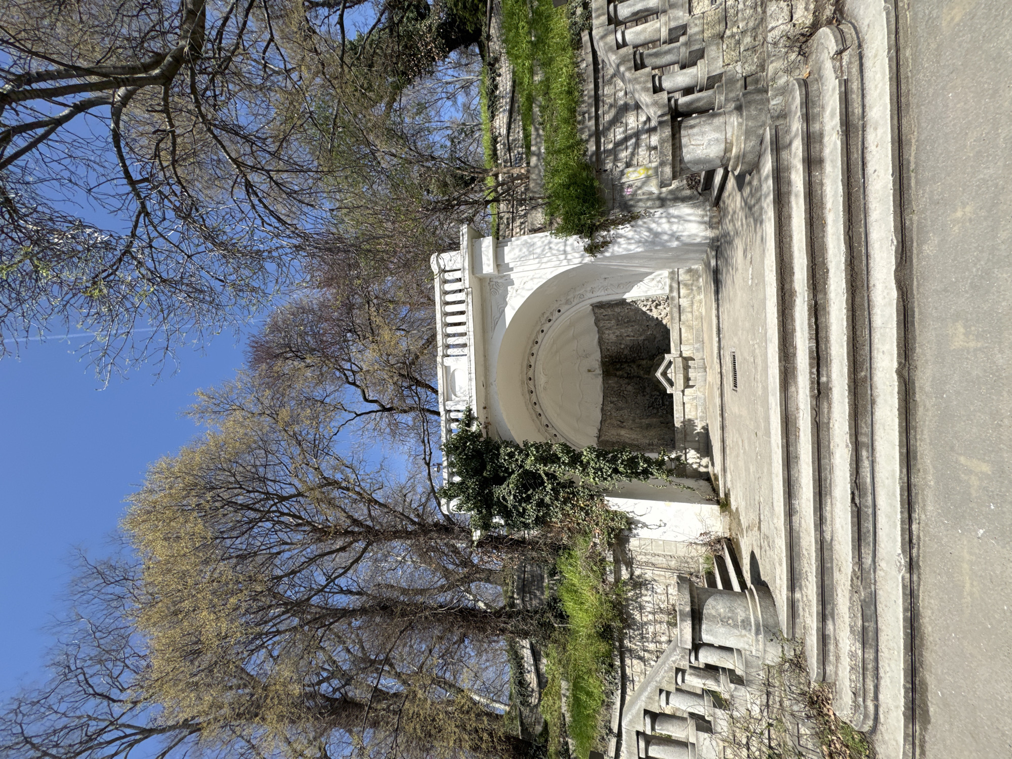 The image captures an outdoor scene dominated by a white, ornate architectural structure, possibly a historical fountain, grotto, or monument, situated within a park or garden environment. This central feature presents a prominent arched opening, intricate detailing, and decorative balustrades on its upper level. Dark green ivy cascades over parts of the white stone, and other vibrant green foliage grows along adjacent walls and steps.
To the right of the main structure, a series of broad, concrete steps or terraces ascend, showing signs of age with worn surfaces and dirt accumulation in their grooves. The background and upper portions of the image are filled with several large deciduous trees. Some of these trees still display bare branches, while others are beginning to show nascent, light green leaves, indicating early spring.
The scene is illuminated by bright daylight under a clear, vivid blue sky, suggesting sunny weather. The presence of strong shadows cast by both the architecture and trees indicates a mid-morning or early afternoon time. No people or specific activities are depicted, creating a peaceful and serene atmosphere. The architectural style, with its curves and classic elements, points to an older, possibly historic, public space. There is no legible text visible within the frame.