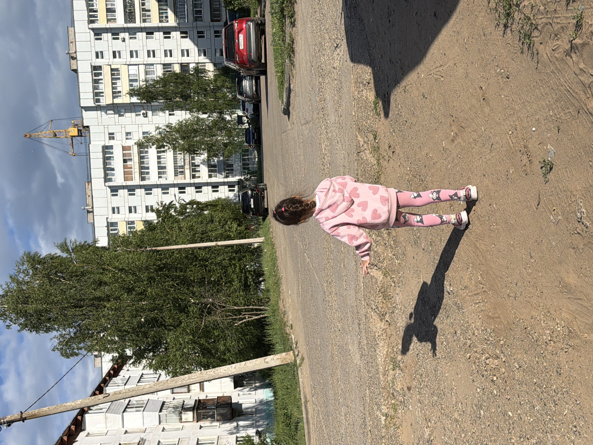 A young girl wearing a pink hoodie with heart patterns and pink leggings with cat illustrations is walking on a dirt path. She has brown hair styled in a braid. Her shadow is cast on the ground beside her. The scene appears to be an urban environment with a large, multi-story building in the background. Trees line the area between the path and the building. There are several parked cars visible along the side of the road. A yellow construction crane is also present near the building. The sky is partly cloudy. The lighting suggests it is daytime, likely late afternoon given the long shadows. There is no visible text in the image. The setting appears to be residential, possibly a courtyard or street in Chaikovsky, Russia. The girl's posture suggests she is walking or playing.