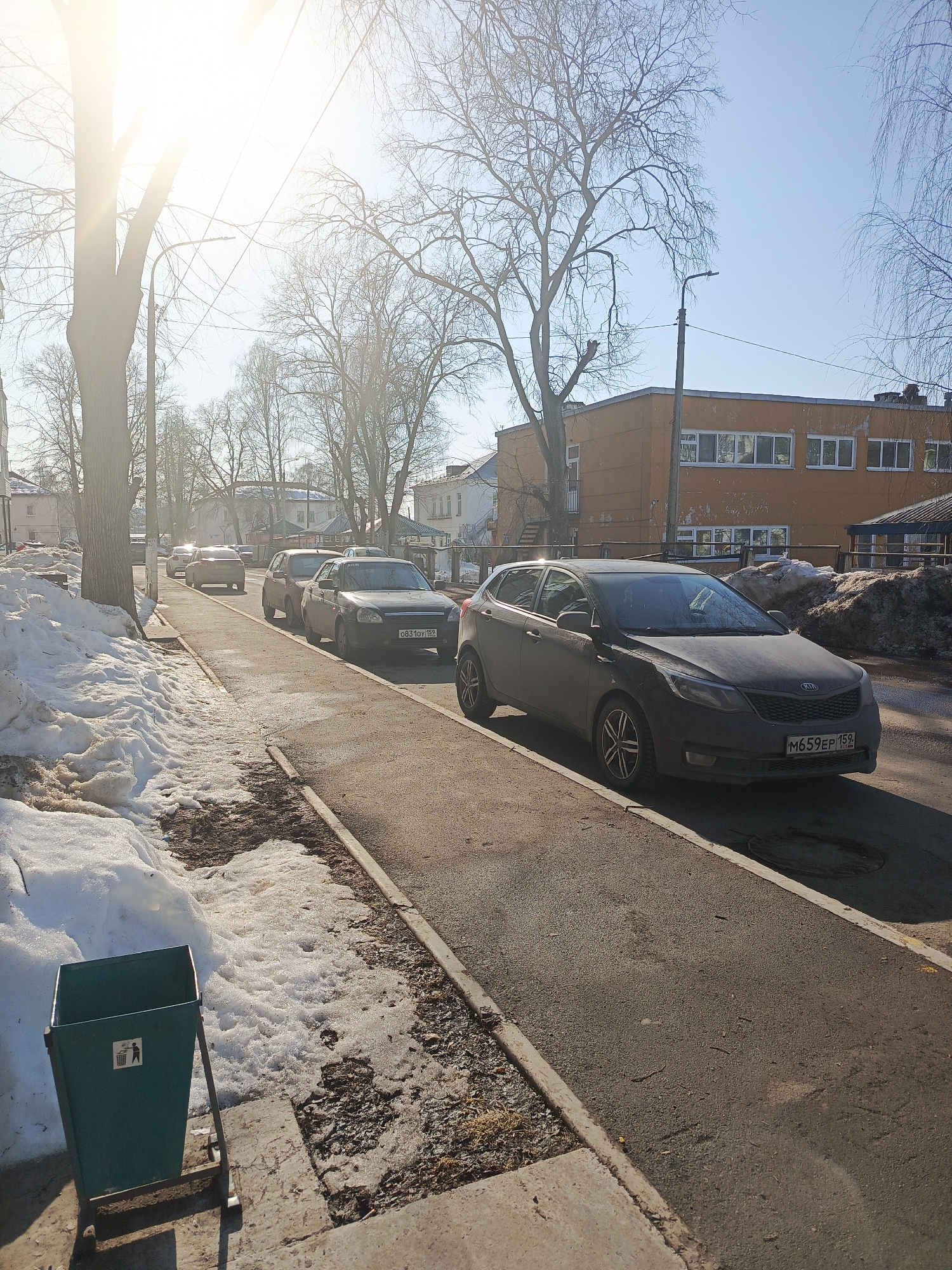 The image depicts a street scene in Chaikovsky, Russia, likely during late winter or early spring, under bright sunlight and a clear blue sky.
The scene is viewed from a slightly elevated perspective on the left side of the street, looking down the thoroughfare. In the immediate foreground on the left, a green metal public trash receptacle with a white pictogram of a person discarding trash stands on a concrete base. Adjacent to it, a curb separates the concrete from an area of melting, dirty snow mixed with exposed soil and bare ground.
To the right of this snow-covered area, an asphalt street extends into the background. A line of vehicles is parked parallel to the curb on the right side of the street. The foremost vehicle is a dark grey Kia Rio hatchback, displaying Russian license plate "M659EP 159". Behind the Kia, a dark grey Lada Priora sedan is parked, bearing Russian license plate "O831OY 159". Further back, a red sedan and additional vehicles are visible.
On the left side of the street, large, bare deciduous trees line the sidewalk, with one prominent tree trunk partially obscuring the intense sunlight originating from the upper left quadrant of the image. This sunlight produces significant lens flare. Overhead, utility wires crisscross the frame.
To the right of the parked vehicles, beyond a curb, another substantial pile of dirty, melting snow is accumulated. Behind this snow pile and the parked cars, an orange-colored, multi-story building with numerous white-framed rectangular windows is visible. A dark street lamp post with an attached luminaire stands in front of this building. No individuals are present in the frame.