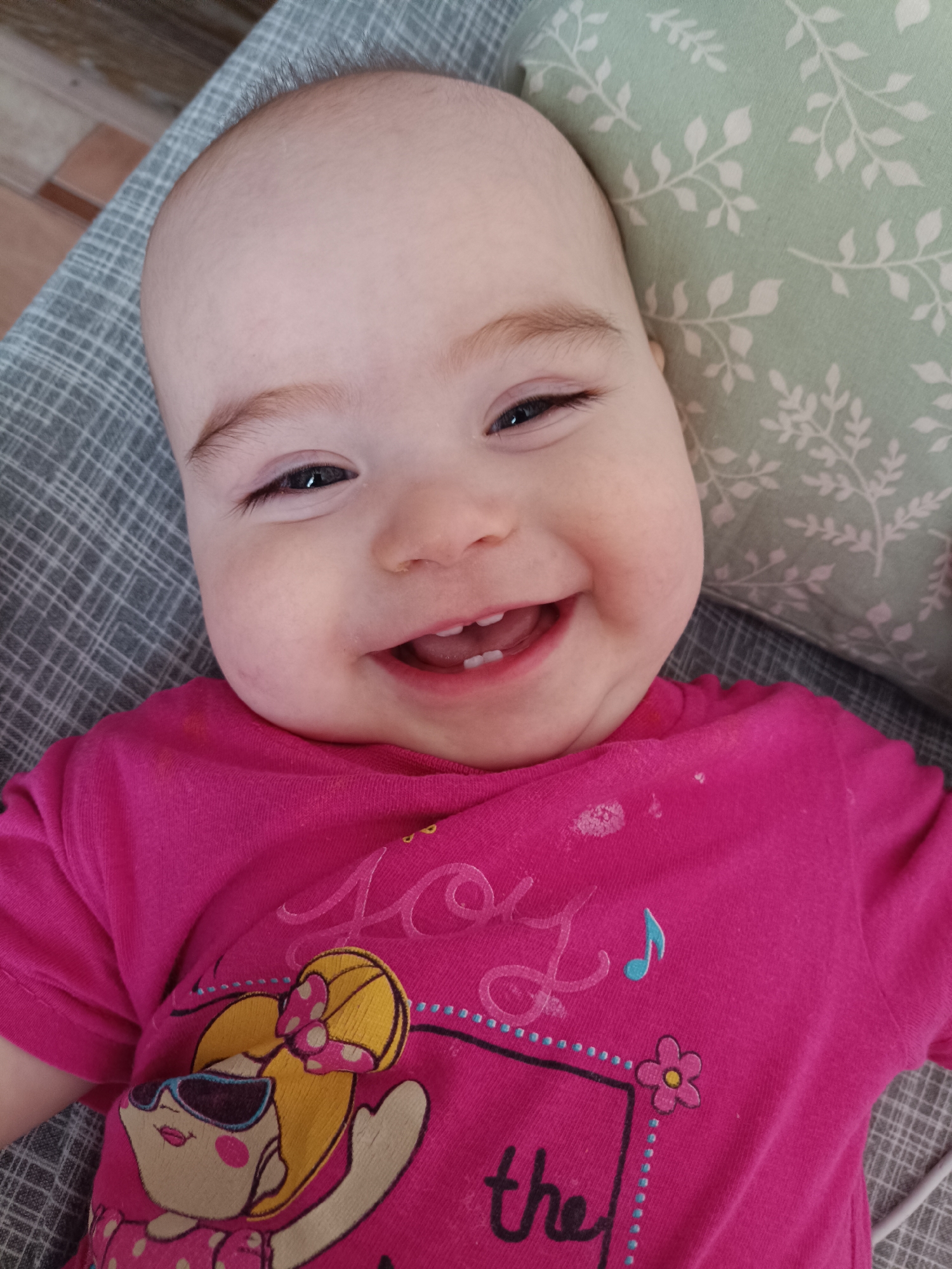 A baby is lying down, smiling at the camera. The baby has a fair complexion, light brown eyebrows, and several teeth visible. The baby is wearing a pink t-shirt with a cartoon design on it. The baby is lying on a gray patterned surface and is partially supported by a green patterned pillow. The location is not discernible from the image.