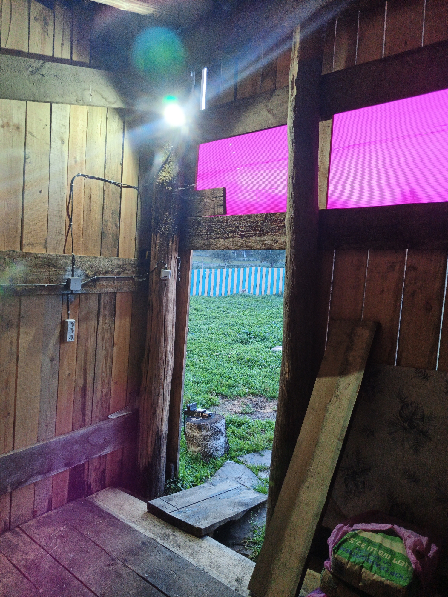 The image depicts the interior of a rustic wooden structure, possibly a shed or stable, in Islambaevo, Russia. The structure is built with rough-hewn wooden planks and beams. A bright LED floodlight is mounted high on the left wall, casting a strong glare and lens flare across the upper portion of the image. Two rectangular panels of translucent pink material are visible in the upper right section of the structure, acting as windows or skylights.
In the foreground, the floor is made of wooden planks. A wooden bench is visible on the left side. A doorway or opening in the center of the structure leads to an outdoor area. Through the opening, a patch of green grass is visible, bordered by a fence with vertical blue and white stripes. A tree stump with some objects on top is situated just outside the opening.
On the left interior wall, electrical conduits and outlets are visible. In the foreground on the right, leaning against the interior wall, is a long, rough wooden plank. Partially visible in the lower right corner is a pink plastic bag containing what appears to be a green bag with text on it. The overall impression is a utilitarian, rural setting.