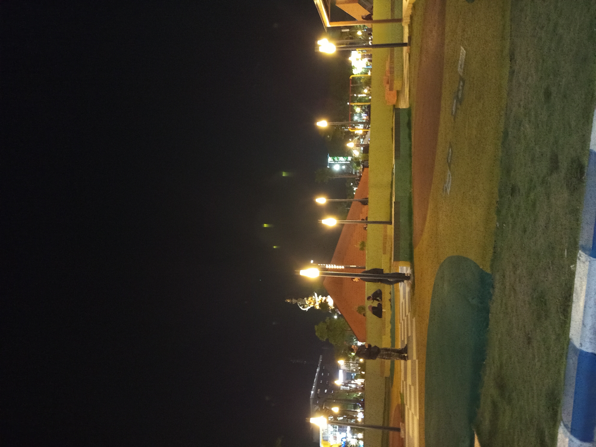 The image shows a nighttime scene in a park or public space in Sidoarjo, Indonesia. In the foreground, a paved area with a checkered pattern is visible, transitioning into a grassy area. Several modern-style lampposts with bright, diffused lights illuminate the scene. Two men are standing and looking at their phones in the mid-ground. Behind them, several other people are seated on a bench. Further in the background, there is a large, conical-shaped structure, possibly a monument or building, with lights at its peak. In the far background, buildings with illuminated windows can be seen. The overall impression is of a well-lit urban recreational area at night.