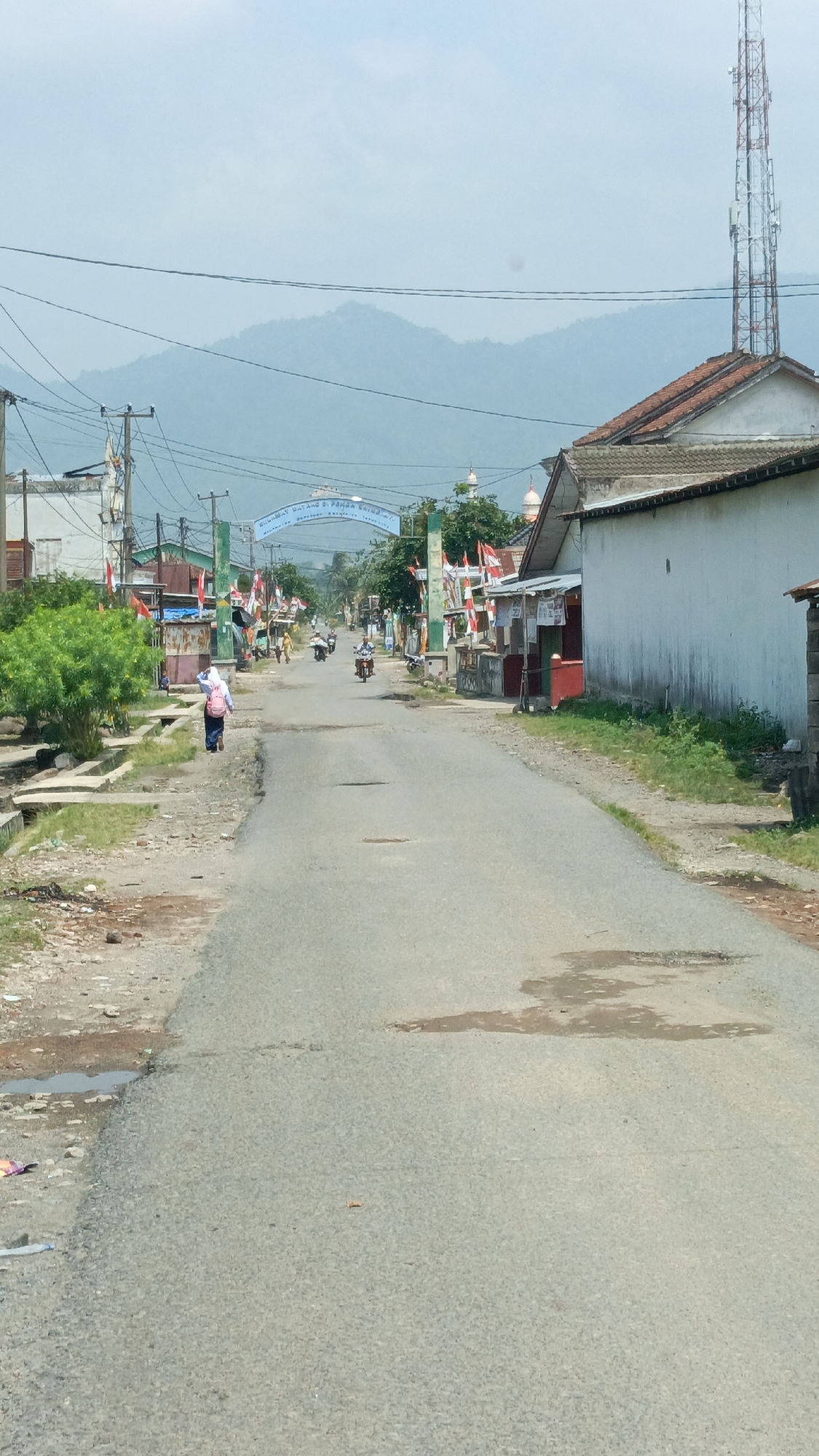 The image depicts a view down a paved road in Kalisari, Indonesia. The road is in moderate condition with some potholes visible. On the left side of the road, there are concrete drainage channels, a low wall, and sparse vegetation. A person in a headscarf and pink backpack is walking away from the camera towards the center of the image. On the right side of the road, there are houses with tiled roofs and white walls. A tall telecommunications tower is visible in the background, as are rolling hills and a clear sky. The road is lined with numerous Indonesian flags, suggesting a celebration or national event. Several motorcycles are present on the road, with at least two people riding them. An archway with text is visible in the distance, likely marking an entrance to a specific area or town. The overall scene is a typical rural or suburban street in Indonesia on what appears to be a clear day.