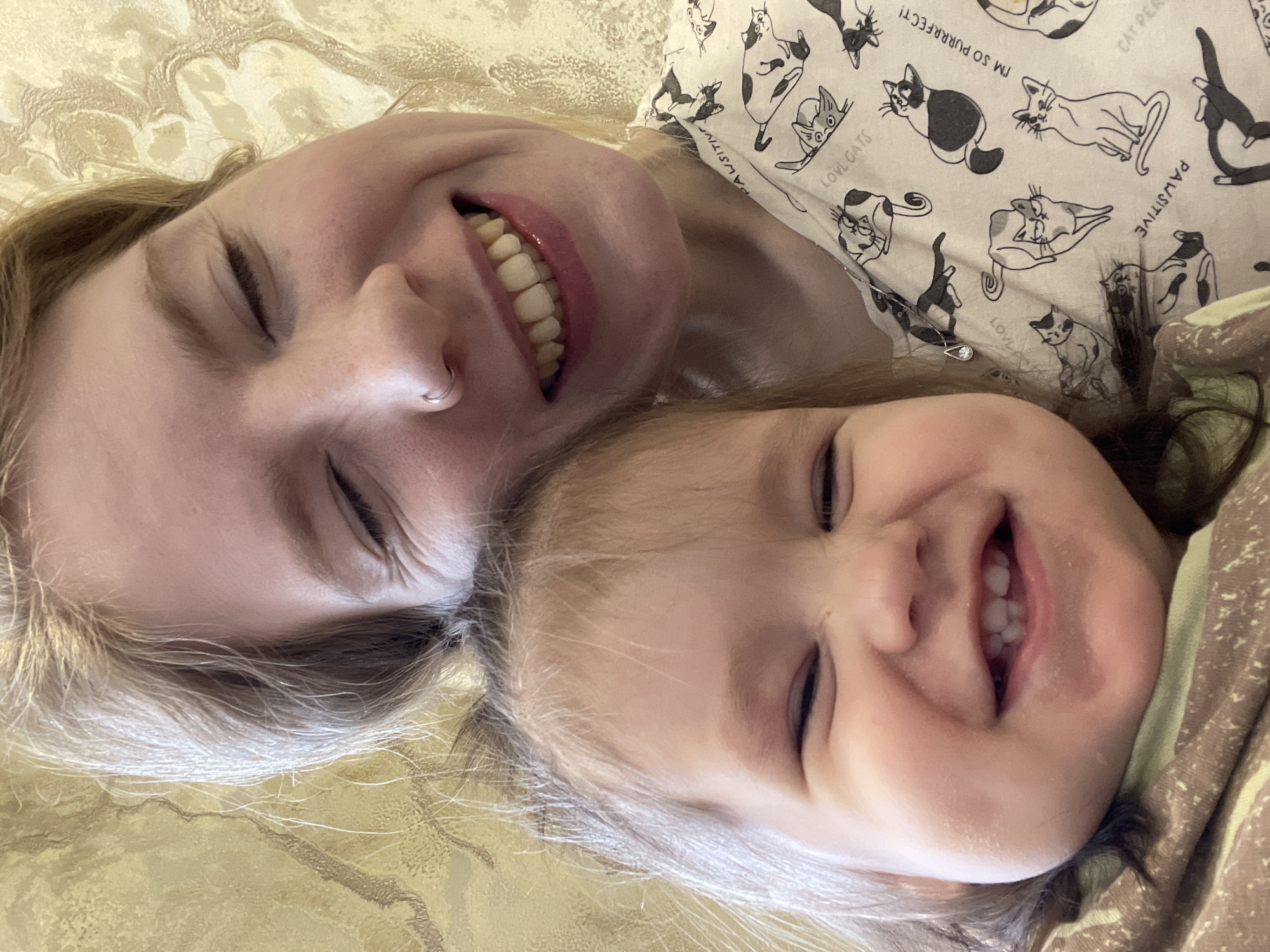Two people, an adult female and a young child, are lying down next to each other. Both individuals are smiling with their eyes closed. The adult female has blonde hair and a nose ring. She is wearing a white shirt with cat-themed designs. The child has light brown hair and is wearing a brown patterned shirt. They appear to be lying on a patterned gold-colored surface, likely a bed or sofa. The location is not discernible from the image. No information suggests that the image was taken in Izhevsk, Russia.