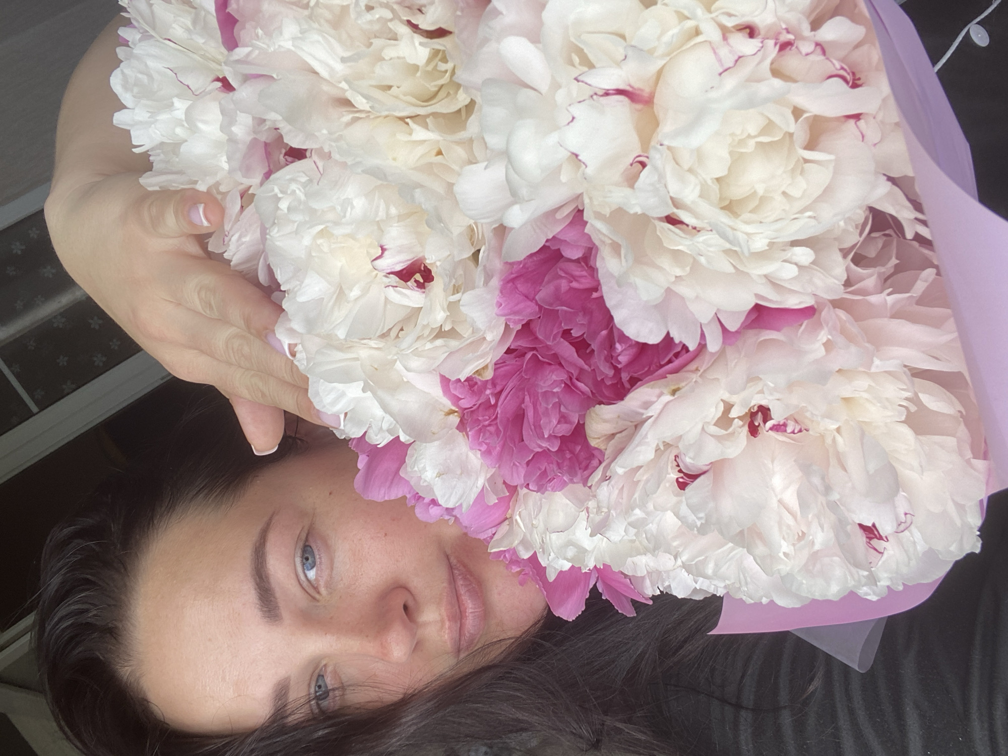 A person is holding a large bouquet of peonies in front of their face. The peonies are primarily white and pink. The person has fair skin, dark hair, and blue eyes. They are lying down, and the bouquet is positioned above them. The background is out of focus, but what can be determined from the background does not appear to be located in Sochi, Russia.
