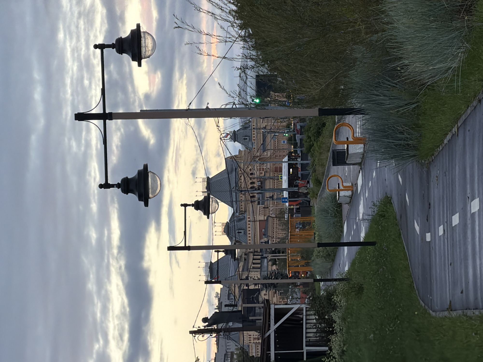 The image depicts a pathway in a park or public space, likely in Nizhny Novgorod, Russia, given the architectural style of the building in the background, which resembles the Fair Building (Nizhegorodskaya Yarmarka). The pathway is paved with asphalt and marked with white lines, suggesting it might be a bike path or a pedestrian walkway with designated lanes. Streetlights with classic lantern-style fixtures are positioned along the pathway.
In the background, a large, multi-story building with intricate roofing, domes, and numerous windows dominates the scene. This building has a distinctly historical and possibly institutional appearance. A statue of a standing figure is visible to the left of the building, positioned on a raised platform.
The environment includes green grassy areas with some ornamental grasses and bushes. In the middle ground, there's a street with a bus and traffic lights. The sky is overcast with dramatic clouds, indicating either early morning or late evening light. The overall atmosphere is calm and somewhat subdued, suggesting a public space during a quiet time of day. There are no visible people interacting.